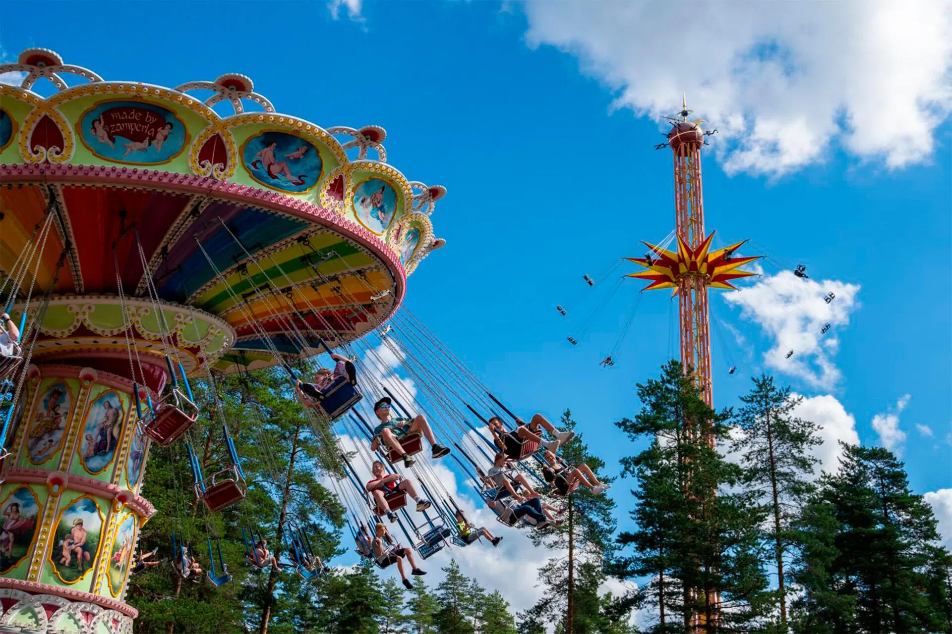 Värikäs ketjukaruselli liikkeessä huvipuistossa, ihmiset keinuvat sinistä taivasta vasten; taustalla korkea tornilaite.