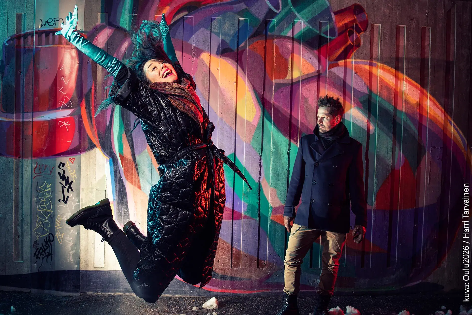 Nainen hyppää ilmaan ja mies katsoo sivulleen värikkäästi maalatun seinän edessä