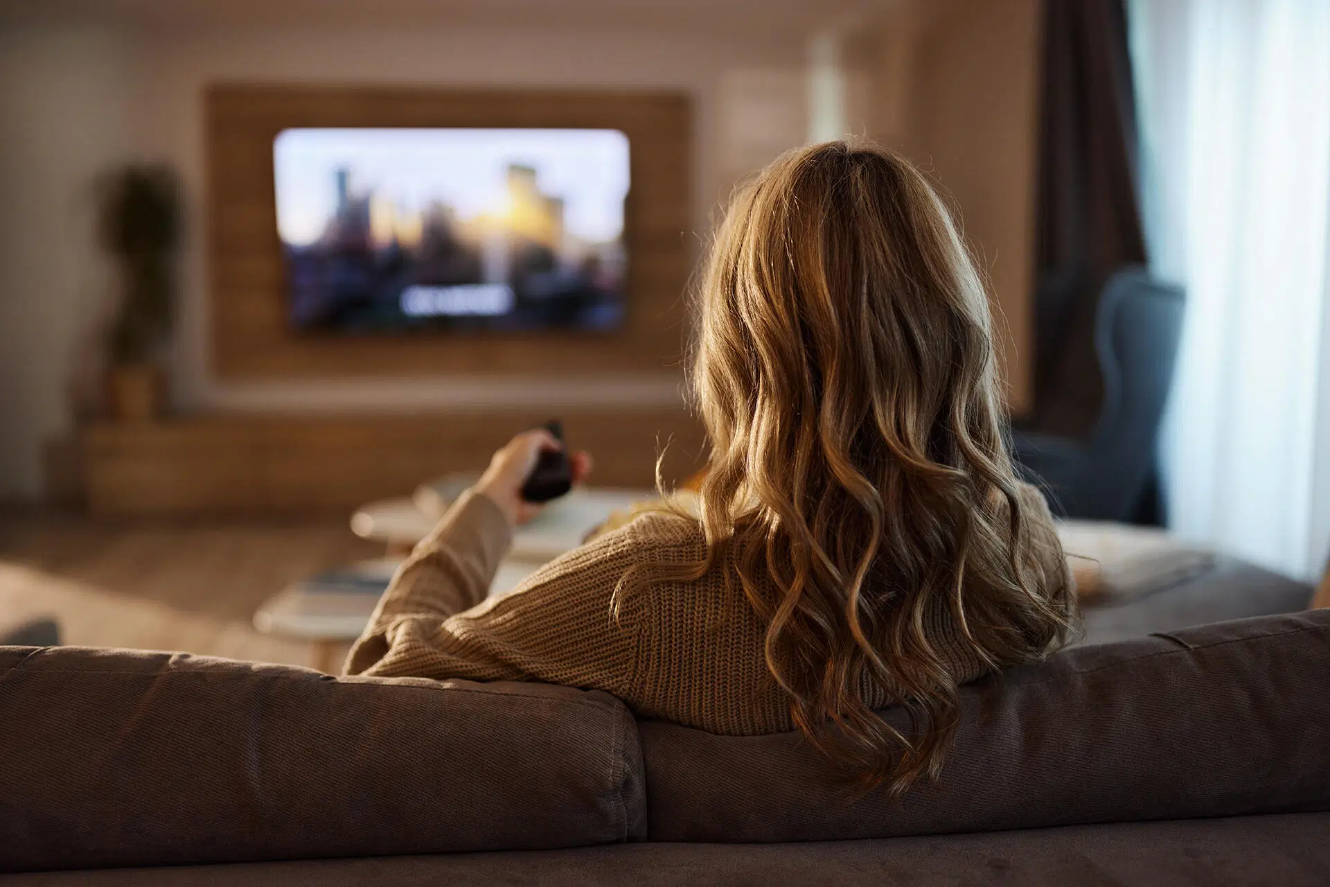 Henkilö sohvalla katsoo televisiota ja pitelee kaukosäädintä olohuoneessa.