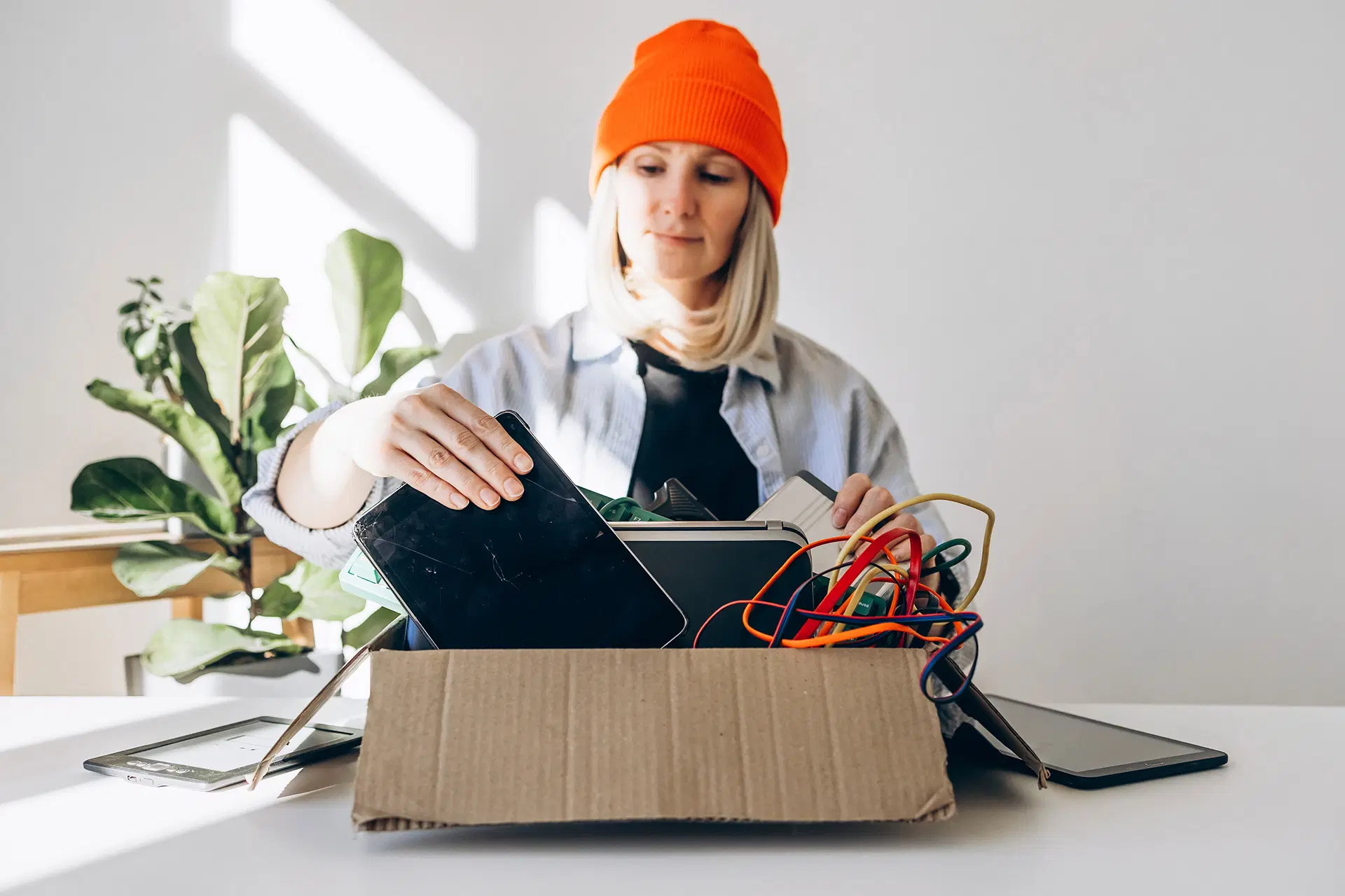 Henkilö oranssissa pipossa lajittelee elektroniikkaa pahvilaatikossa kirkkaassa valossa.