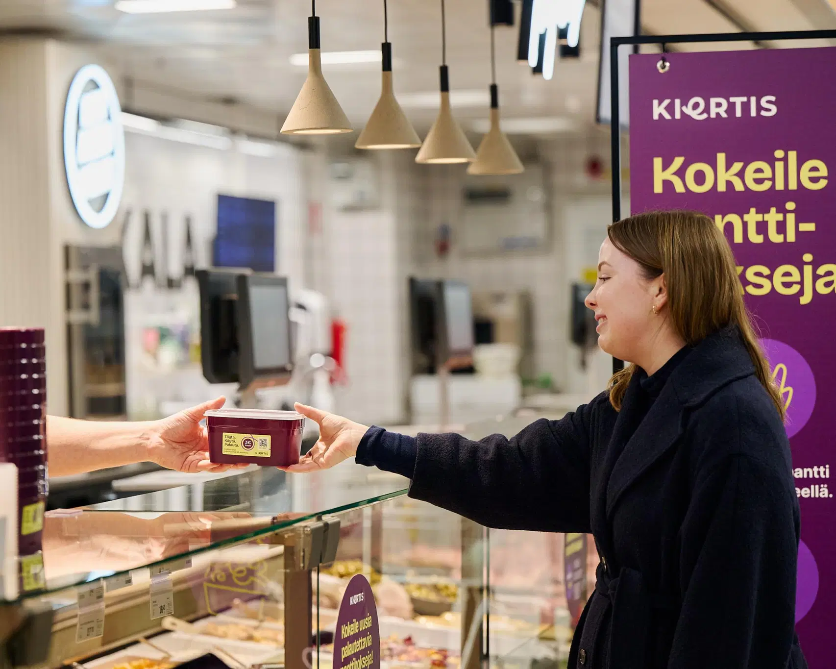 Nainen ottaa vastaan myyjän ojentaman noutoruokarasian kaupan tuoretiskillä.