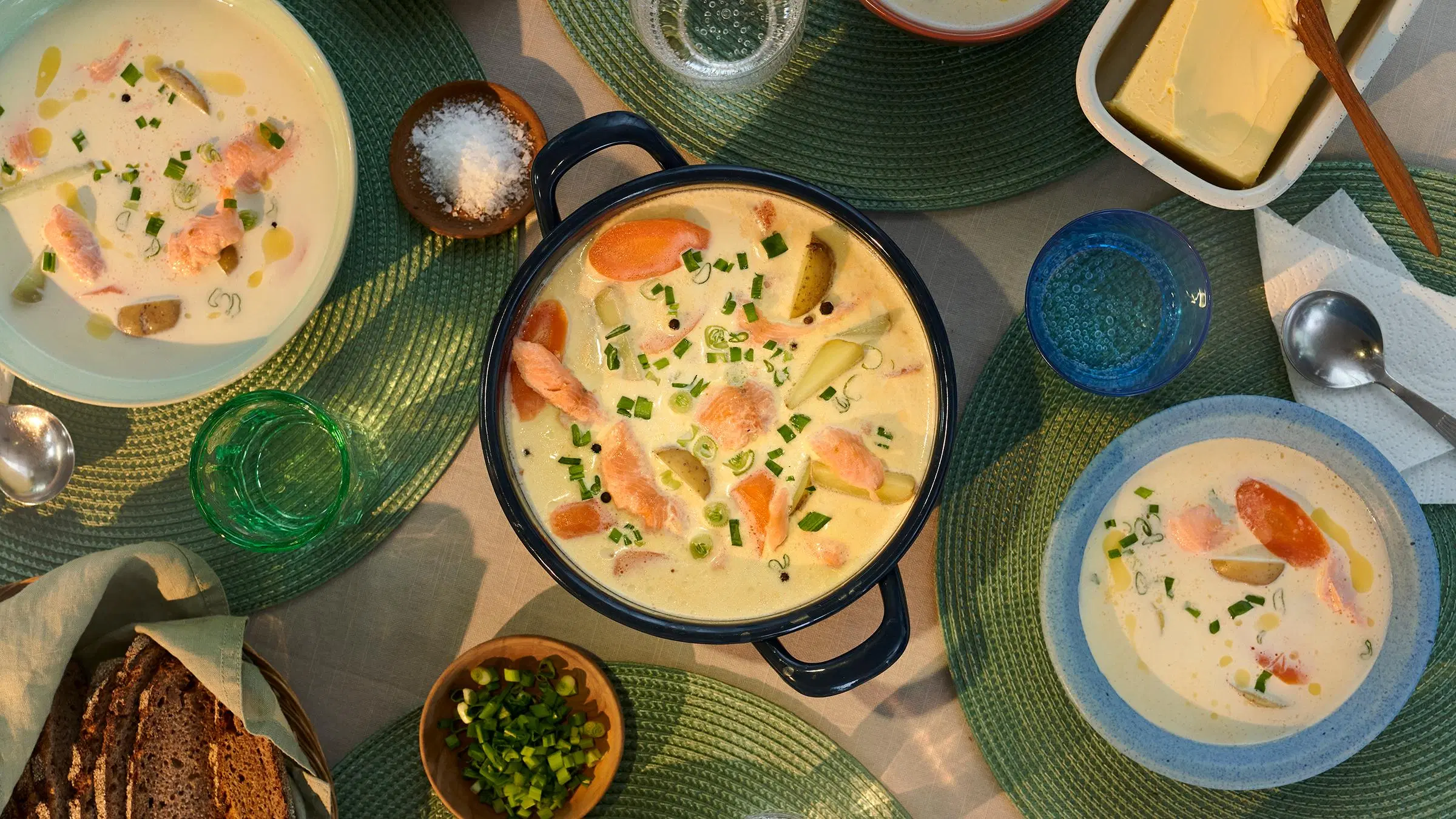 Kermaista savulohikeittoa kattilassa. Vieressä kaksi keittolautasta, leipää, voita ja juomalaseja.