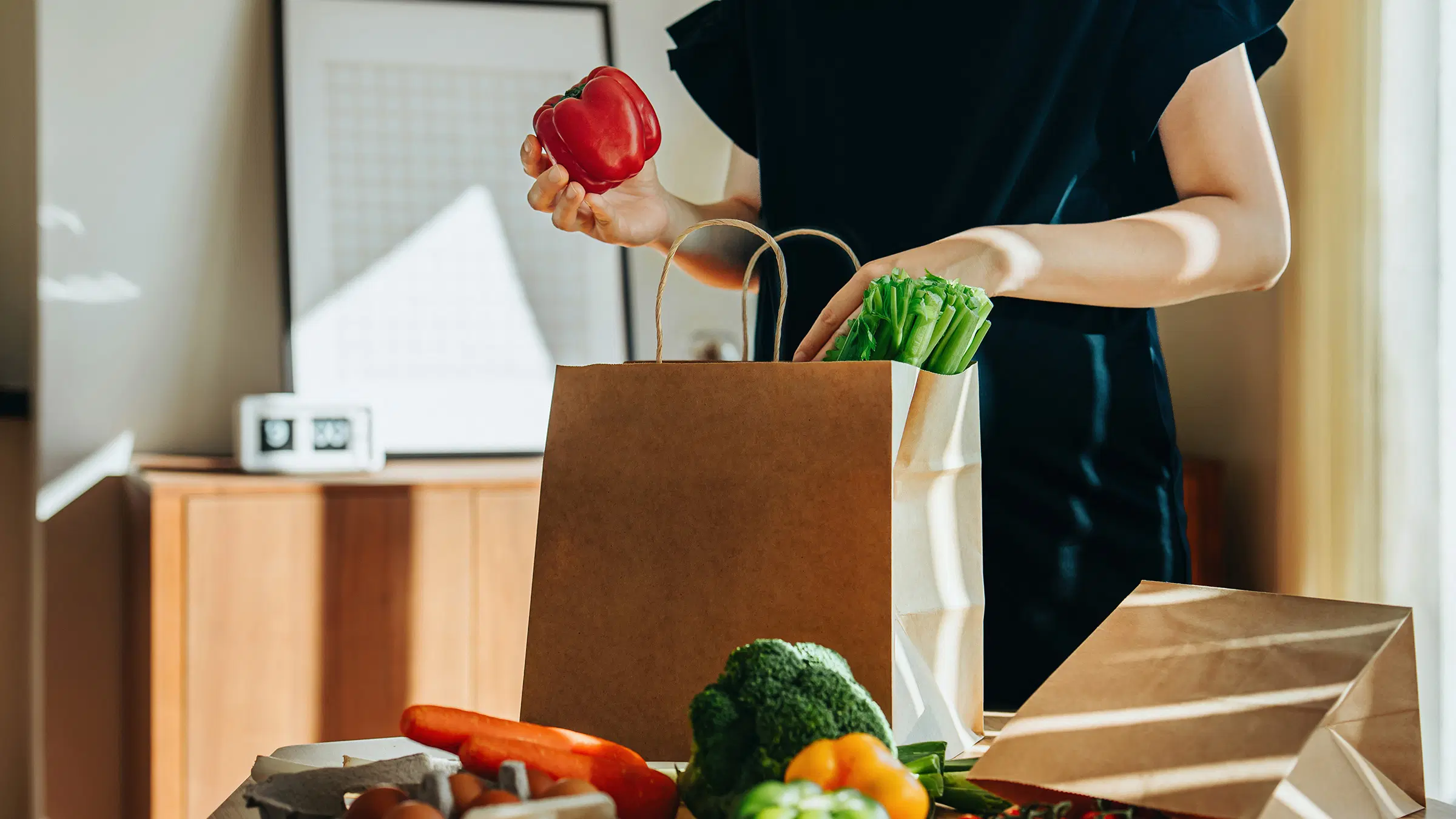 Mustamekkoinen nainen purkaa ruokaostoksia paperikassista.