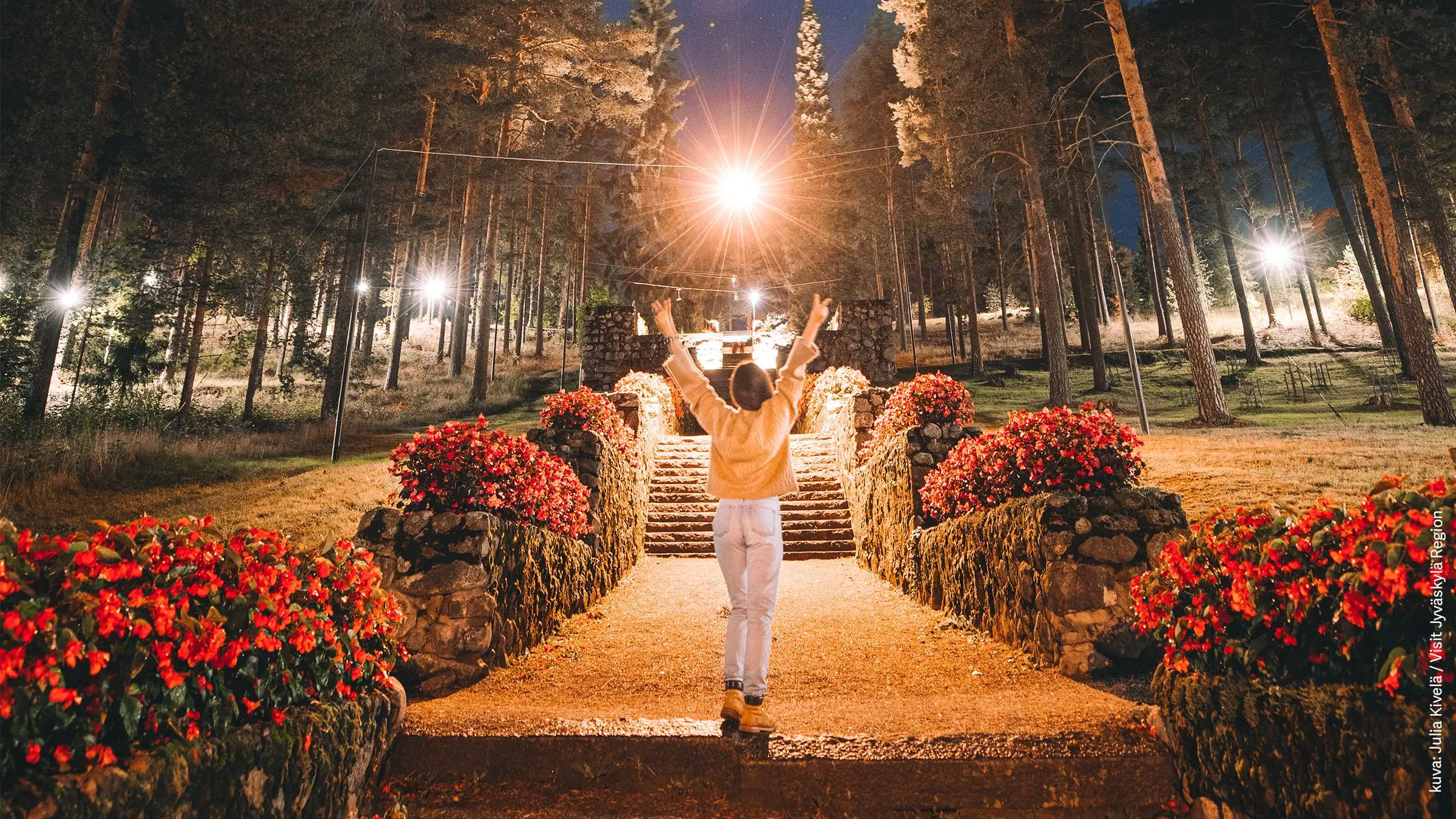 Nuori nainen seisoo iltahämärässä selin kameraan kädet ylhäällä Harjun portailla.