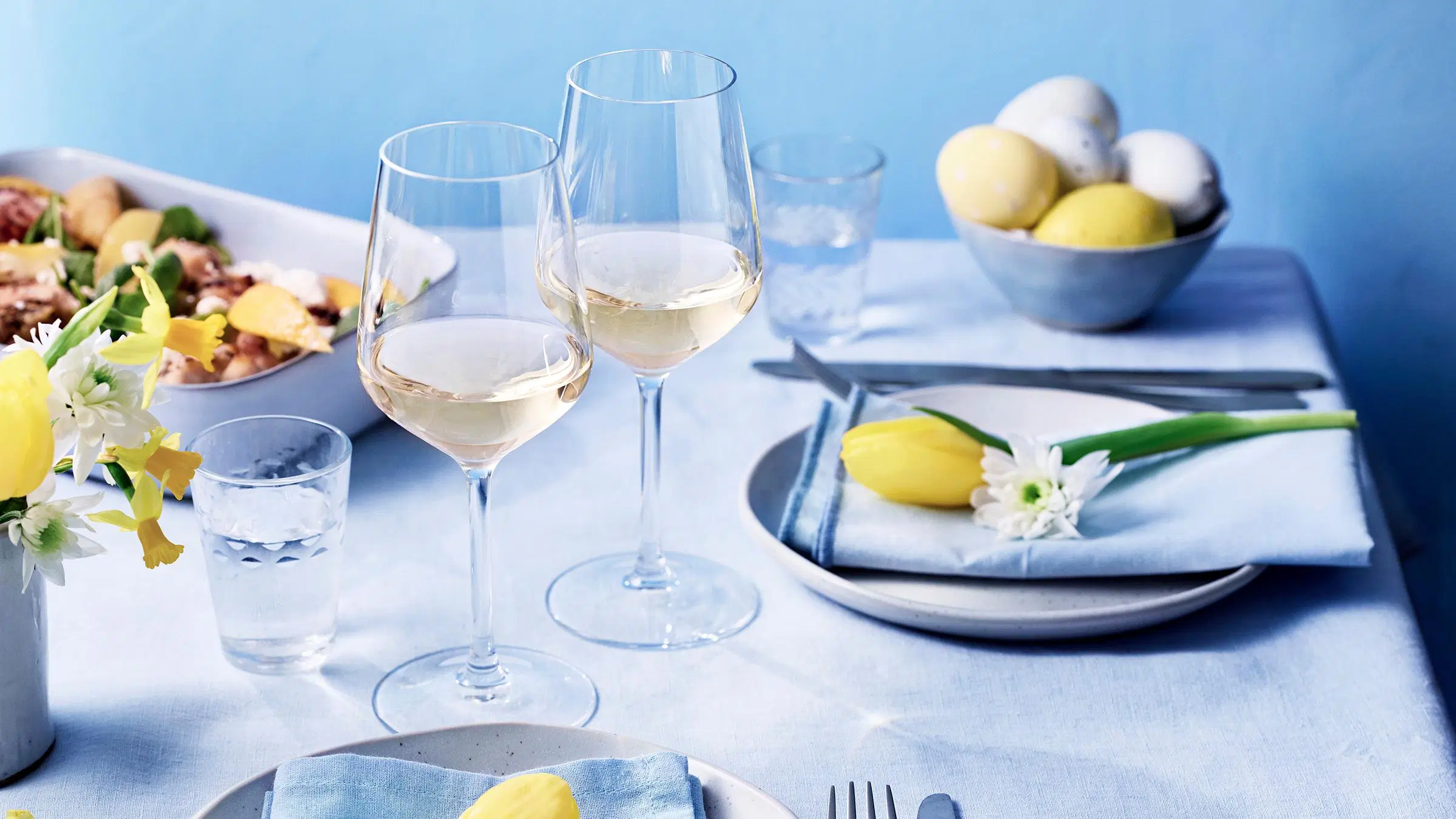 Pääsiäispöydän kattaus, jossa viinilaseja, kukkia, värikkäitä pääsiäismunia ja ruokaa vuoassa