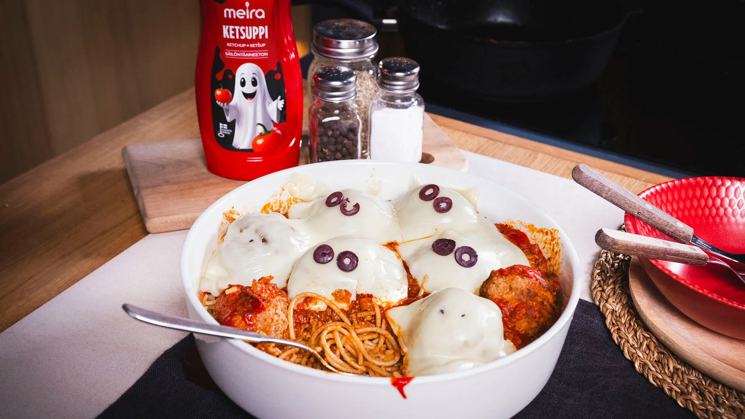 Spagettivuoka lihapullilla, päällä haamuiksi muotoiltua juustoa, joilla oliiviviipaleet silminä, taustalla Meira-ketsuppipullo ja mausteita.