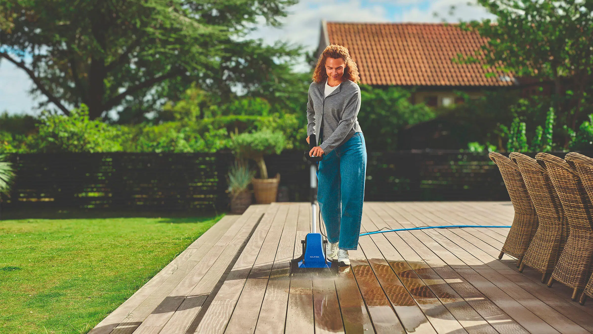 Nainen pesee terassia sinisellä terassipesurilla, taustalla vehreää pihaa