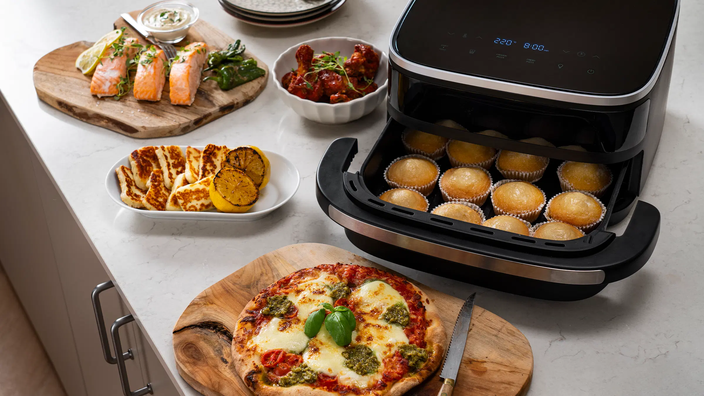 OBH Nordican airfryer paistaa muffineja keittiötasolla; ympärillä pizza, lohta ja muita lämpimiä ruokia.