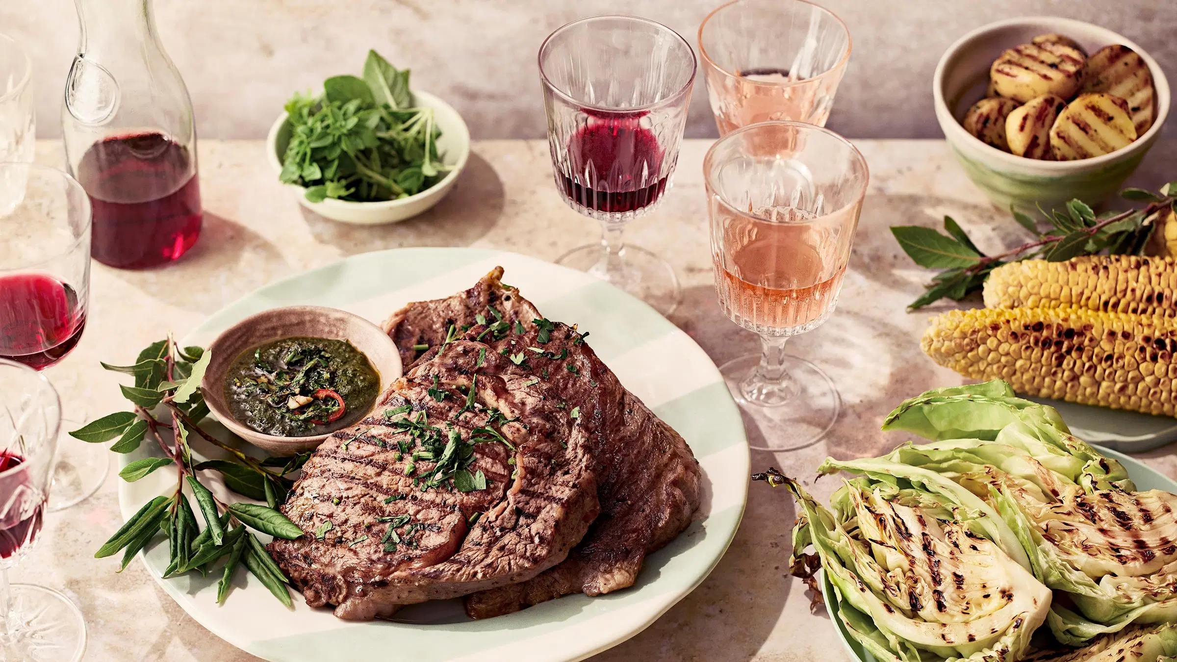 Lautasilla grillattuja pihvejä, maisseja, kaalia ja nektariineja, laseissa juomia.