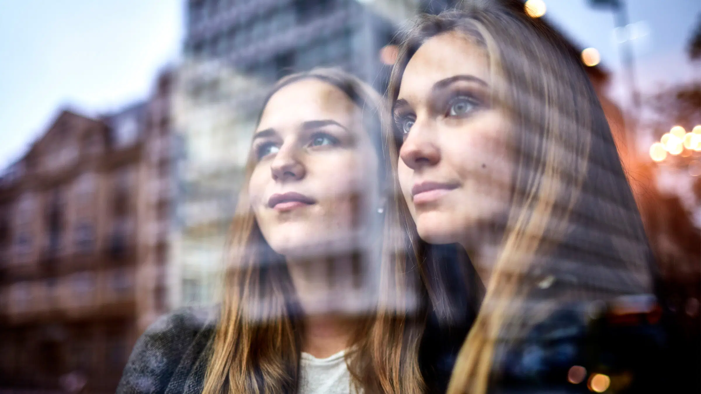 Aikuinen nainen ja nuori nainen lasisen seinän eri puolilla. Lasiin heijastuu kaupungin rakennuksia.