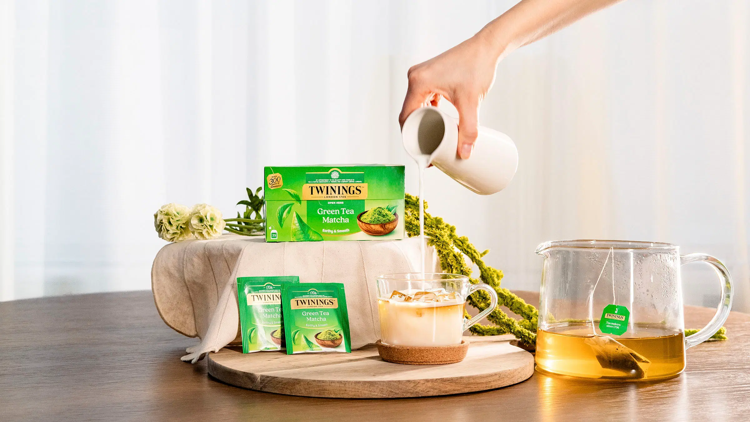 Käsi kaataa kannusta maitoa lasiseen teekuppiin, lasinen teepannu, jossa Twinings-teepussi, Twinings Matcha Green Tea -tuotepakkaus ja kukkia