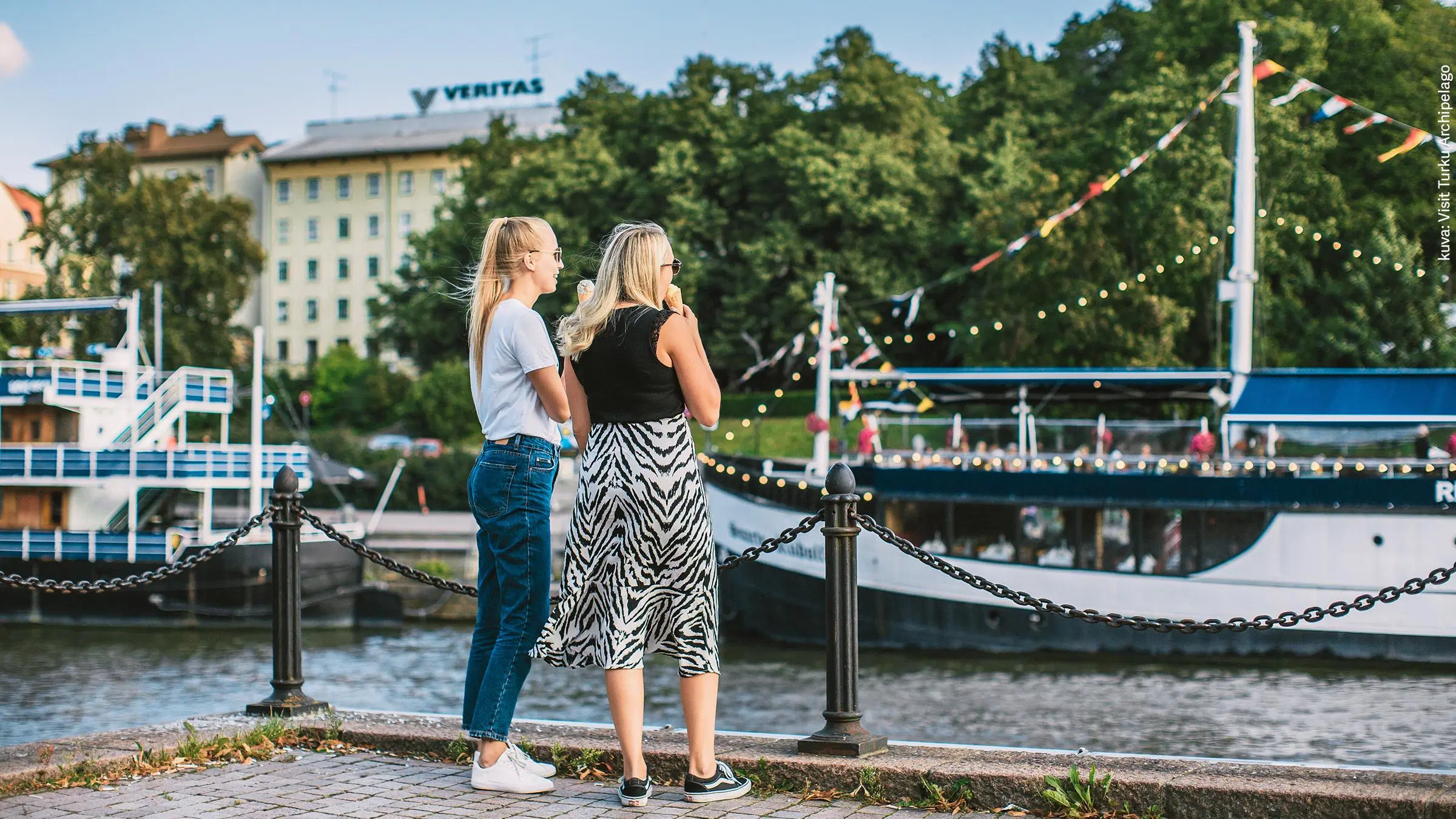 Kaksi takaapäin kuvattua nuorta naista syö jäätelöä joen rannassa. Taustalla jokilaiva.