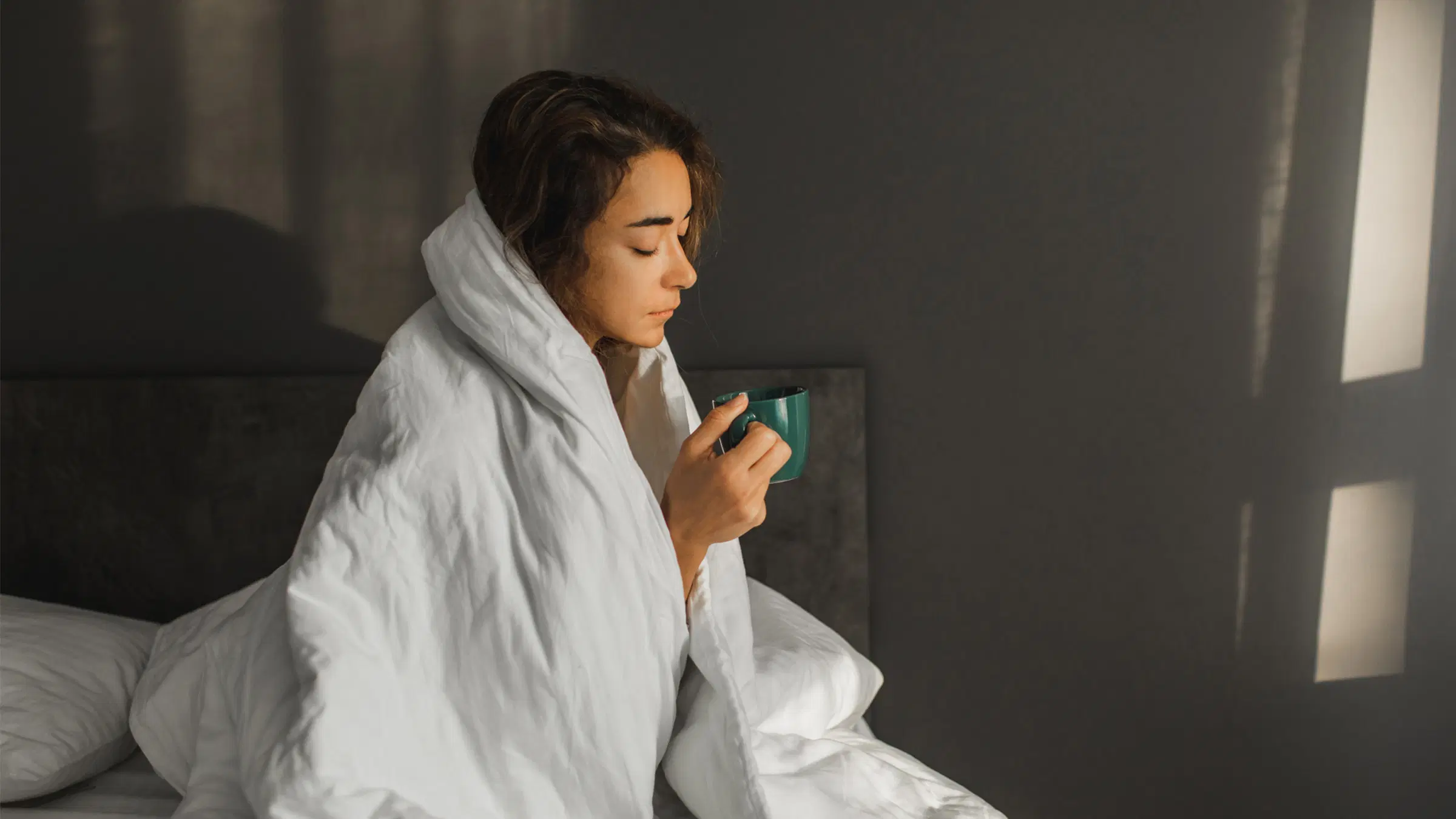 Nainen valkoisen peiton alla kipeän näköisenä vihreä muki kädessä.