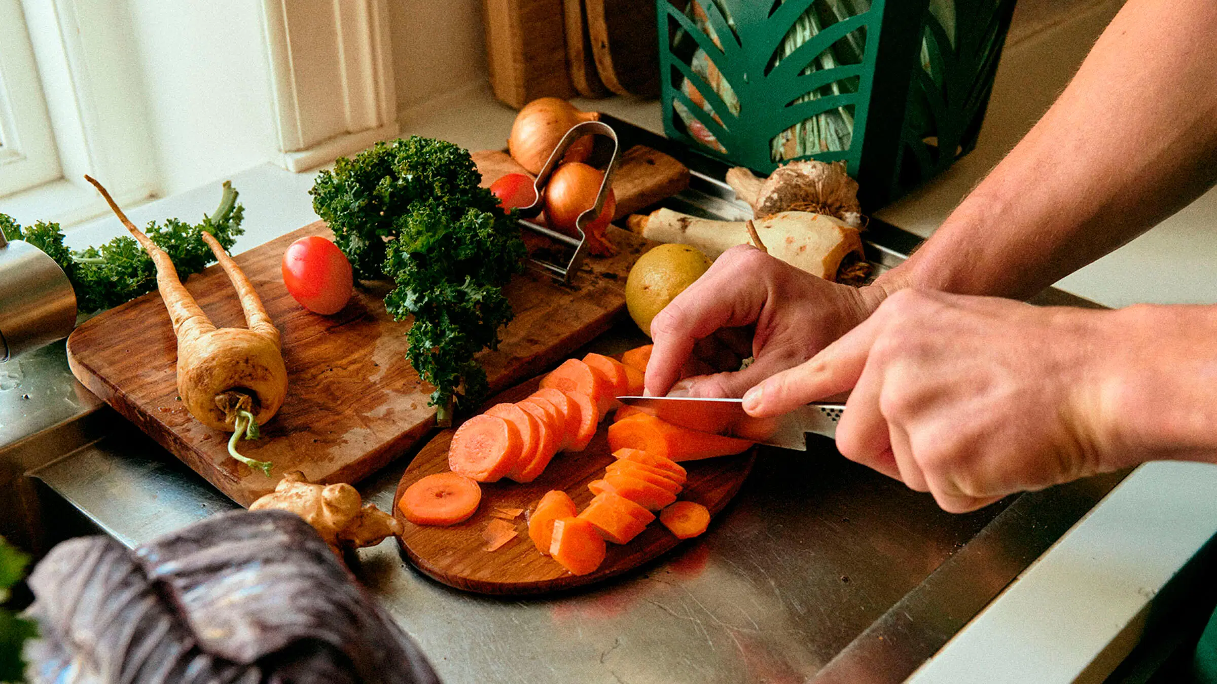 Kädet viipaloivat porkkanaa puisella leikkuulaudalla, ympärillä muita kasviksia ja taustalla biojätesanko.