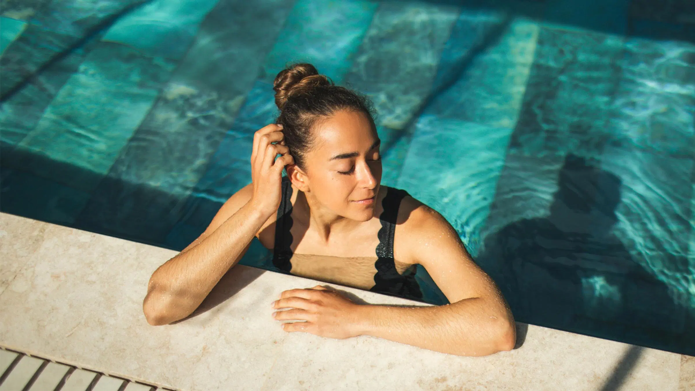 Nainen uima-altaan reunaalla silmät kiinni auringonpaisteessa.