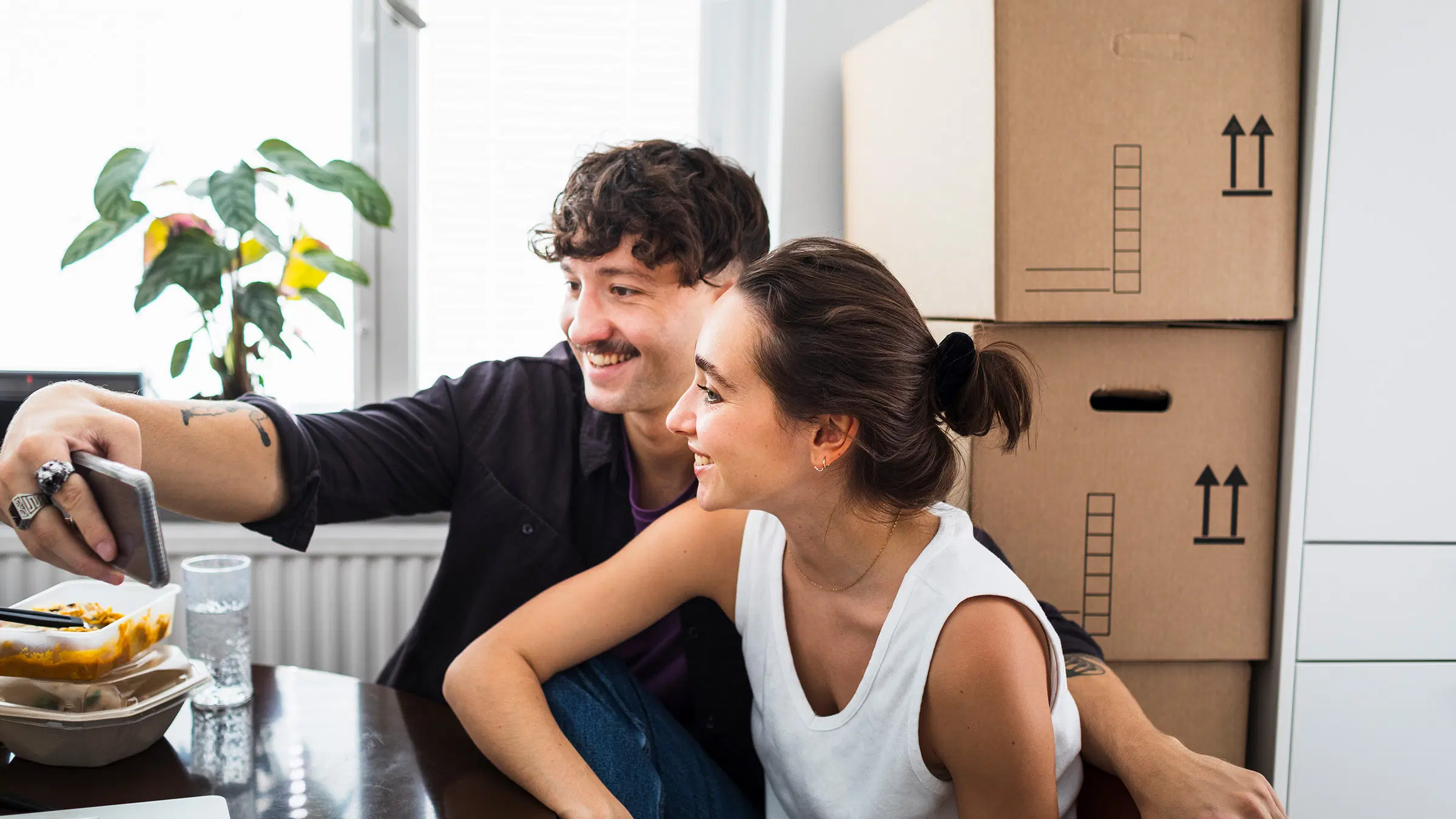 Nainen ja mies katsovat yhdessä puhelinta, takana muuttolaatikoita, pöydällä ruokaa.