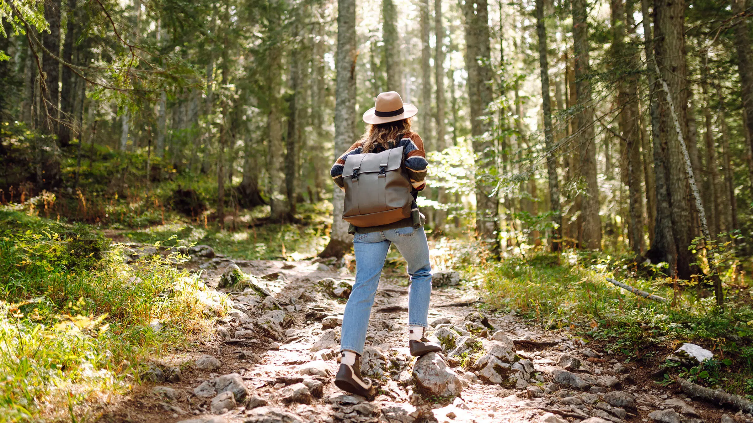 Hattupäinen nainen kulkee metsässä paljasjalkakengissä ja reppu selässä.