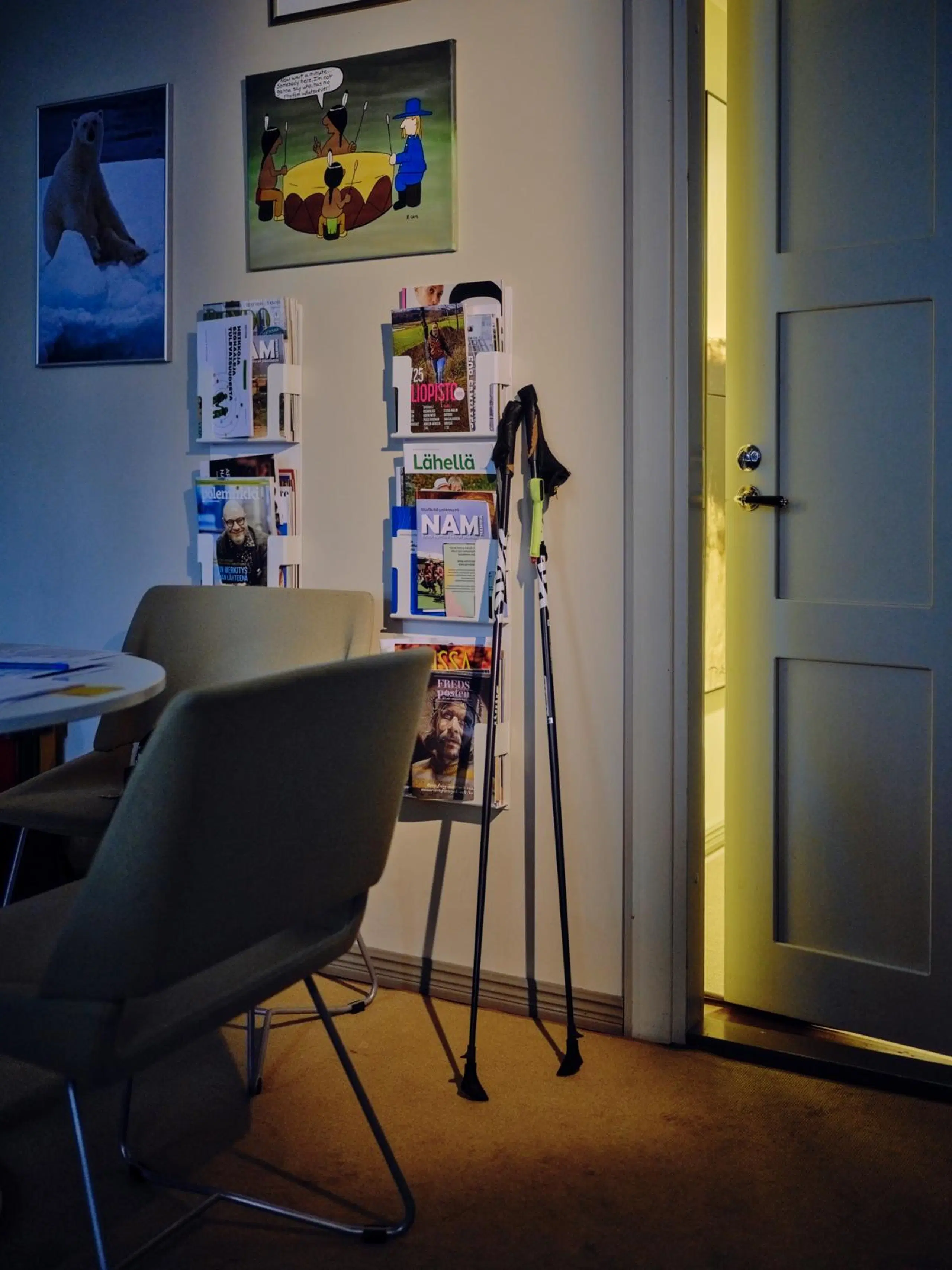 Kävelysauvat nojaavat seinään raollaan olevan oven pielessä. Sauvojen vieressä seinällä on lehtitelineitä ja tauluja.