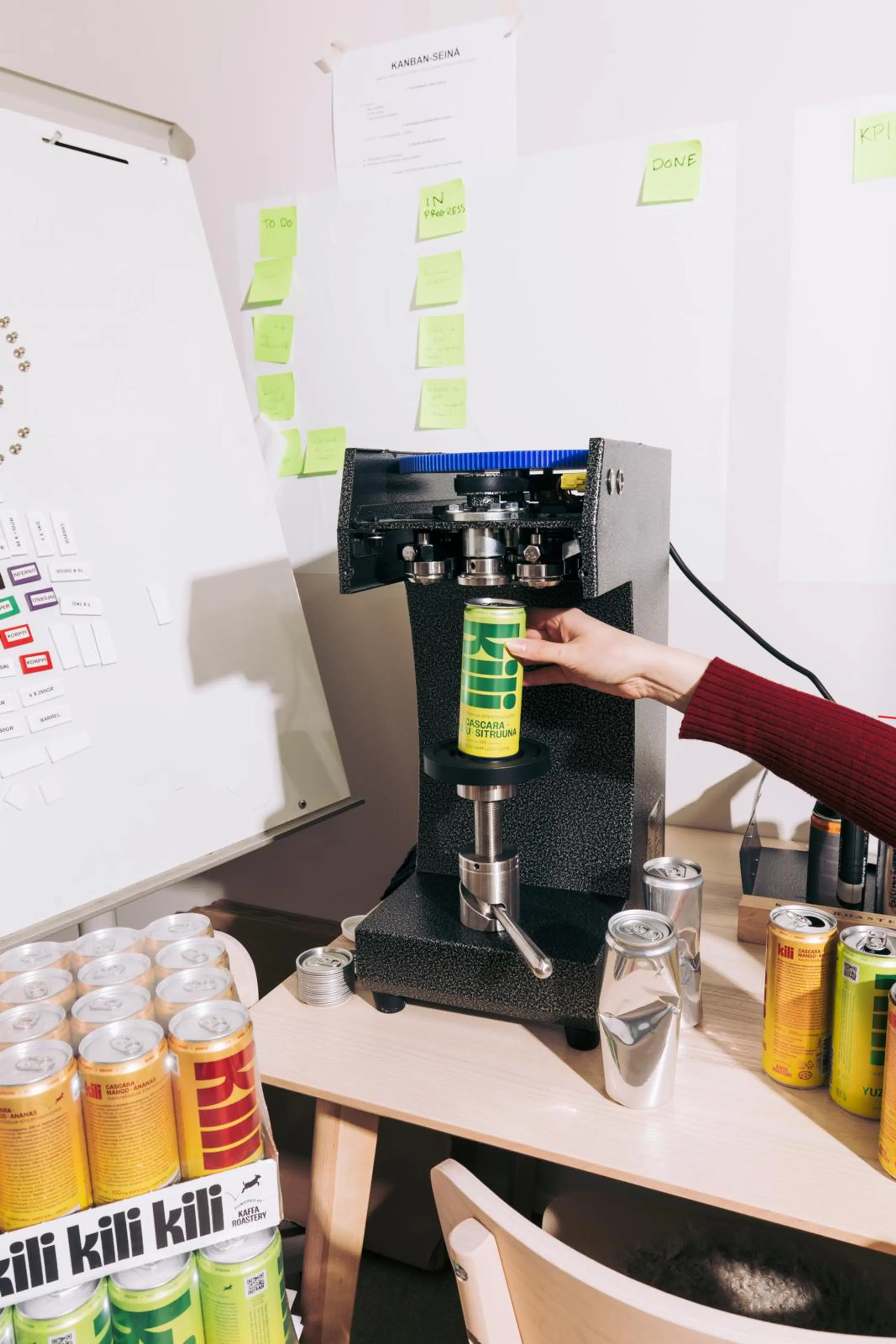 Käsi asettaa Kili-tölkkiä laitteeseen. Ympärillä on lisää Kili-tölkkejä ja kaksi juomatölkkiä ilman etikettejä.