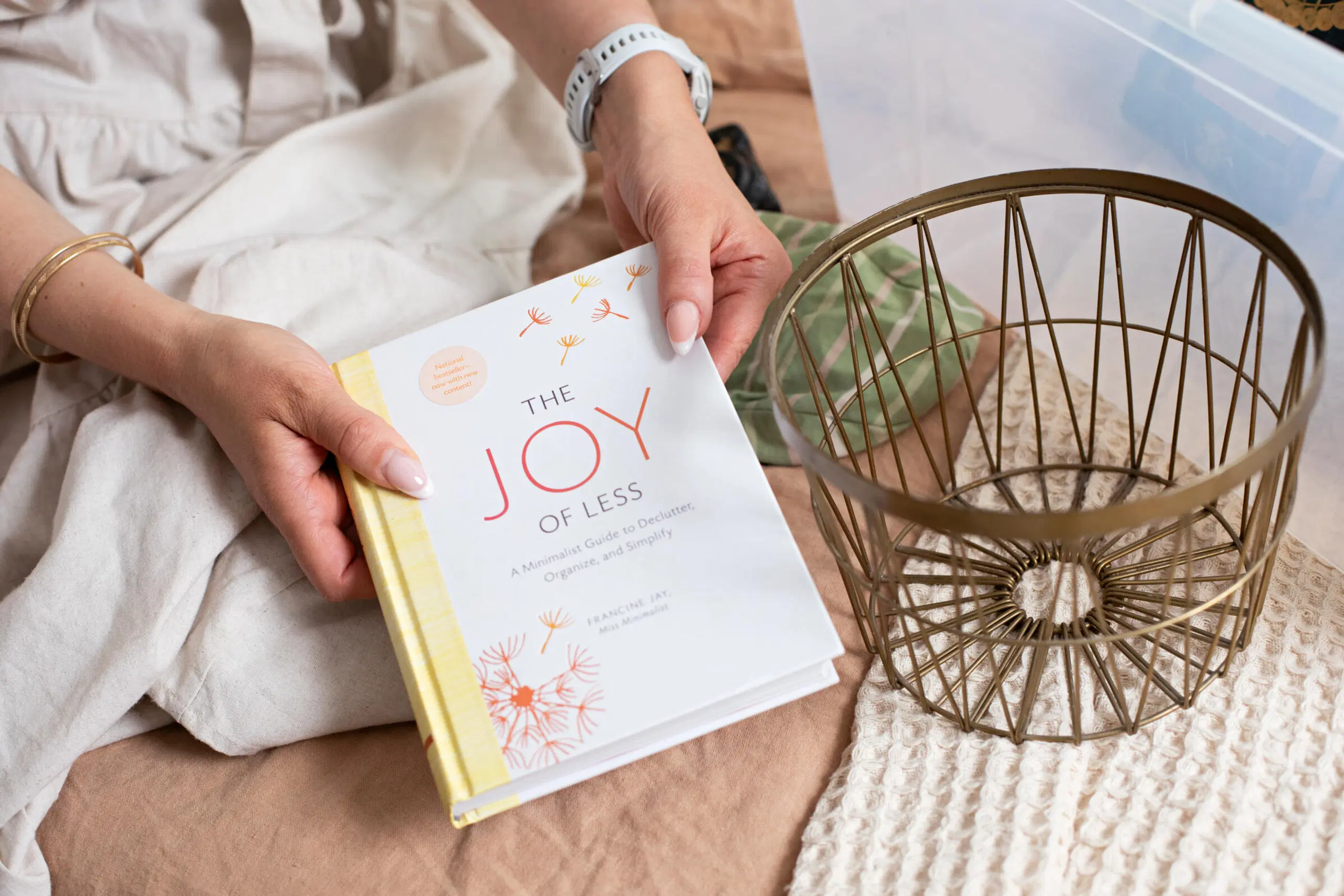 Kädet pitelevät “The Joy of Less” -kirjaa sängyllä, vieressä metallinen kori ja muovilaatikko.