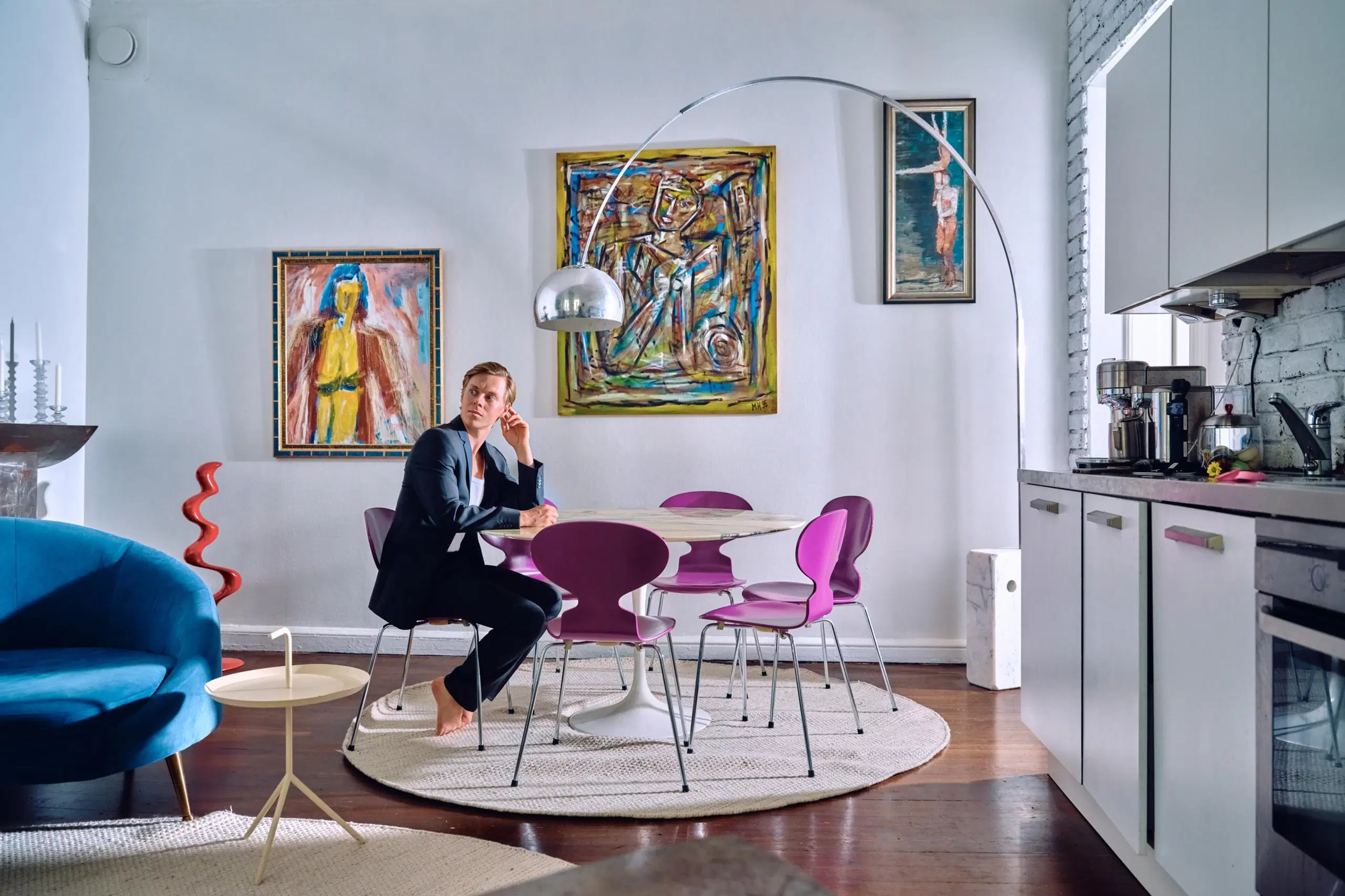 Atte Kilpinen istuu pyöreän ruokapöydän äärellä. Huoneessa on myös krominen kaareva jalkavalaisin ja värikkäitä taideteoksia..