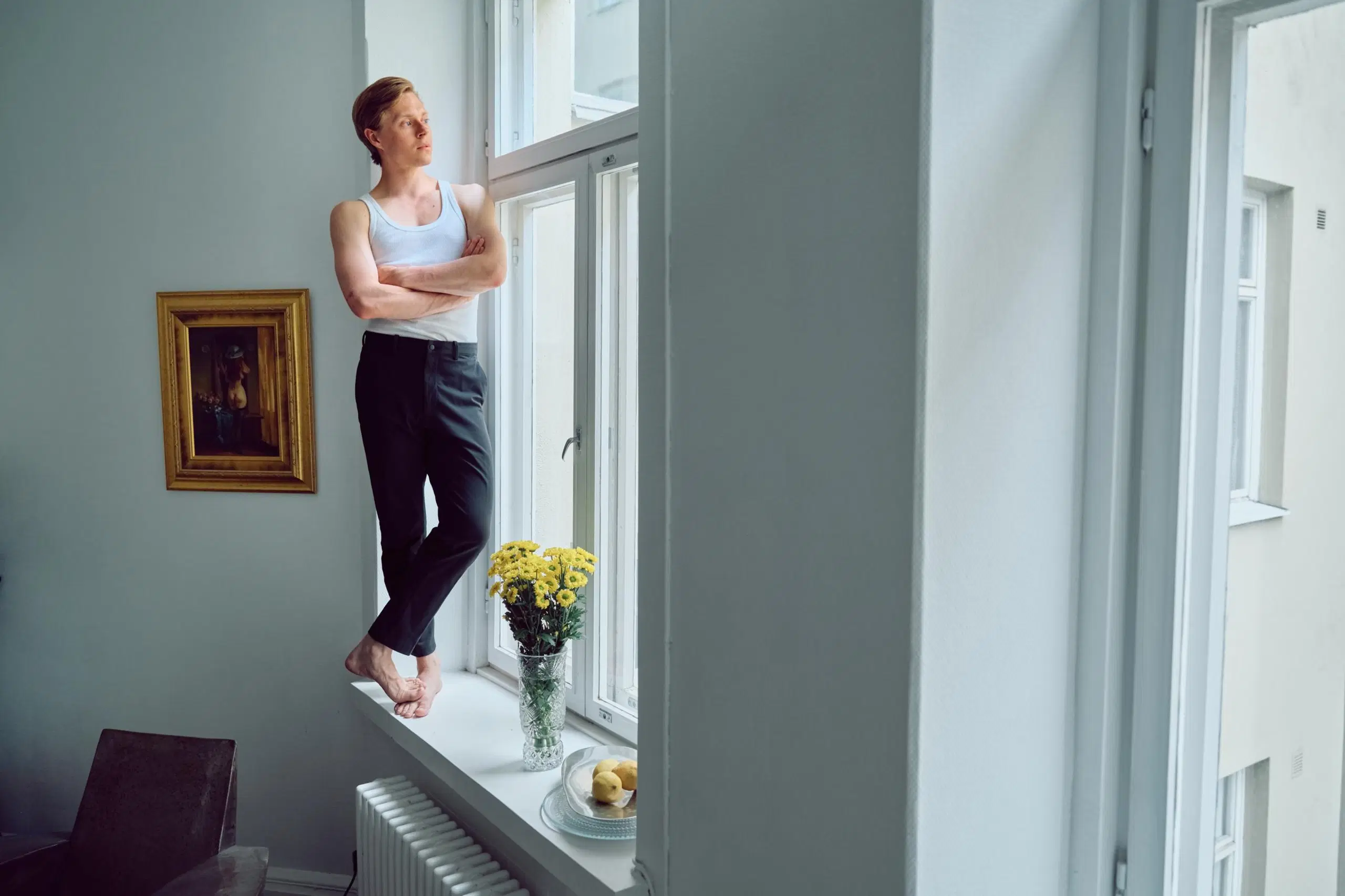 Ate Kilpinen seisoo ikkunalaudalla ja katsoo ulos. Viereisellä seinällä on taulu, joka esittää takaapäin kuvattua vähäpukeista naista.