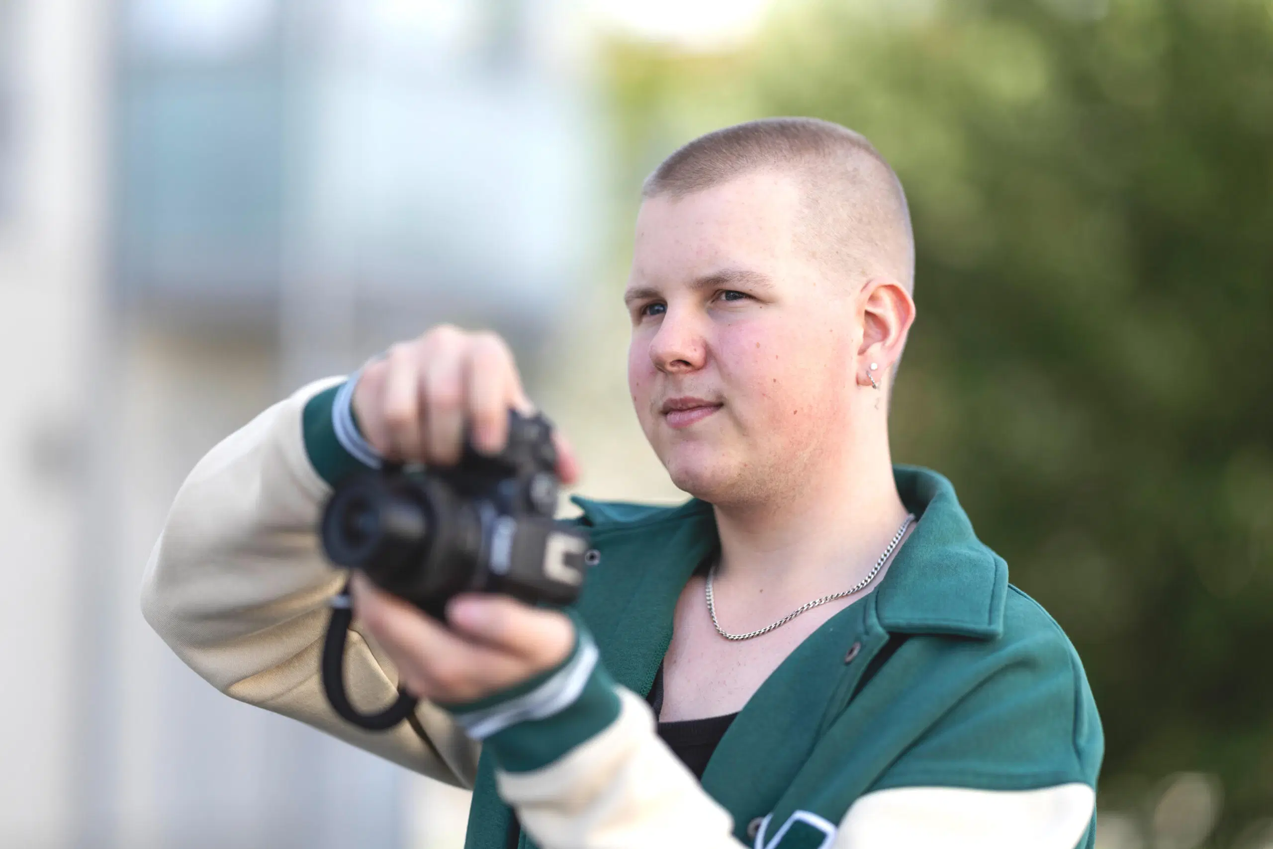 Henkilö ulkona pitelee kameraa ja tähtää kuvaa, vihreä takki, pehmeä tausta.