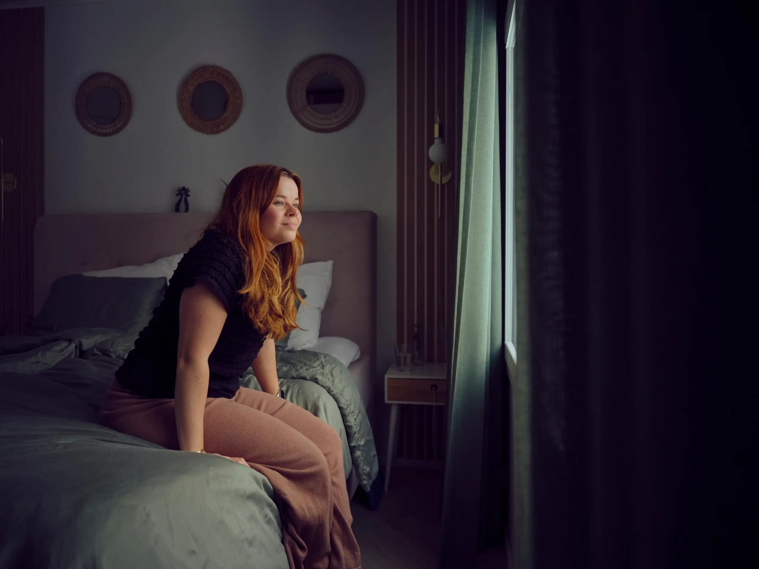 Salla Salmela istuu sängyn reunalla vihreäsävyisessä makuuhuoneessa.