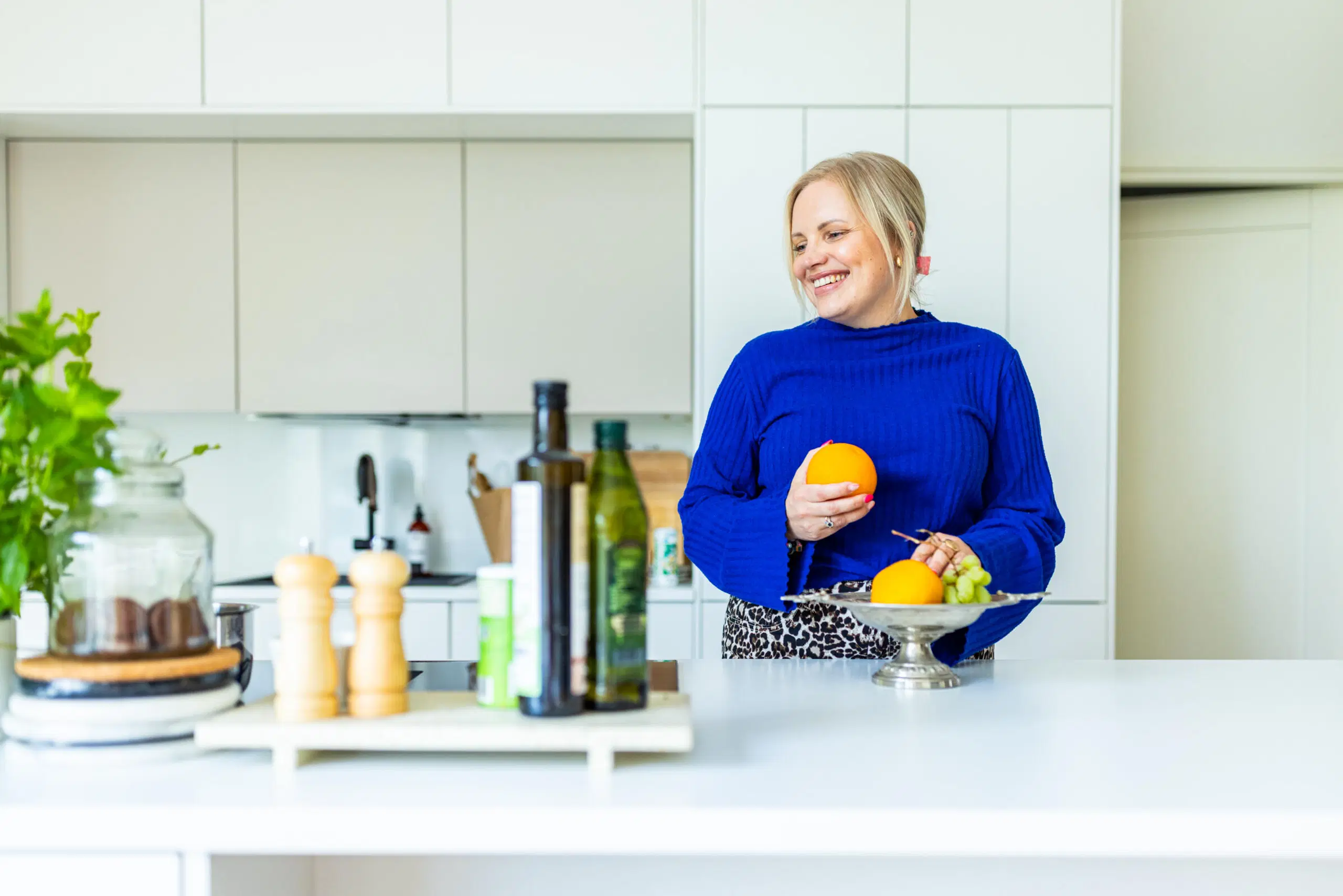 Nainen sinisessä neuleessa hymyilee modernissa keittiössä ja pitää appelsiinia hedelmävadin äärellä.