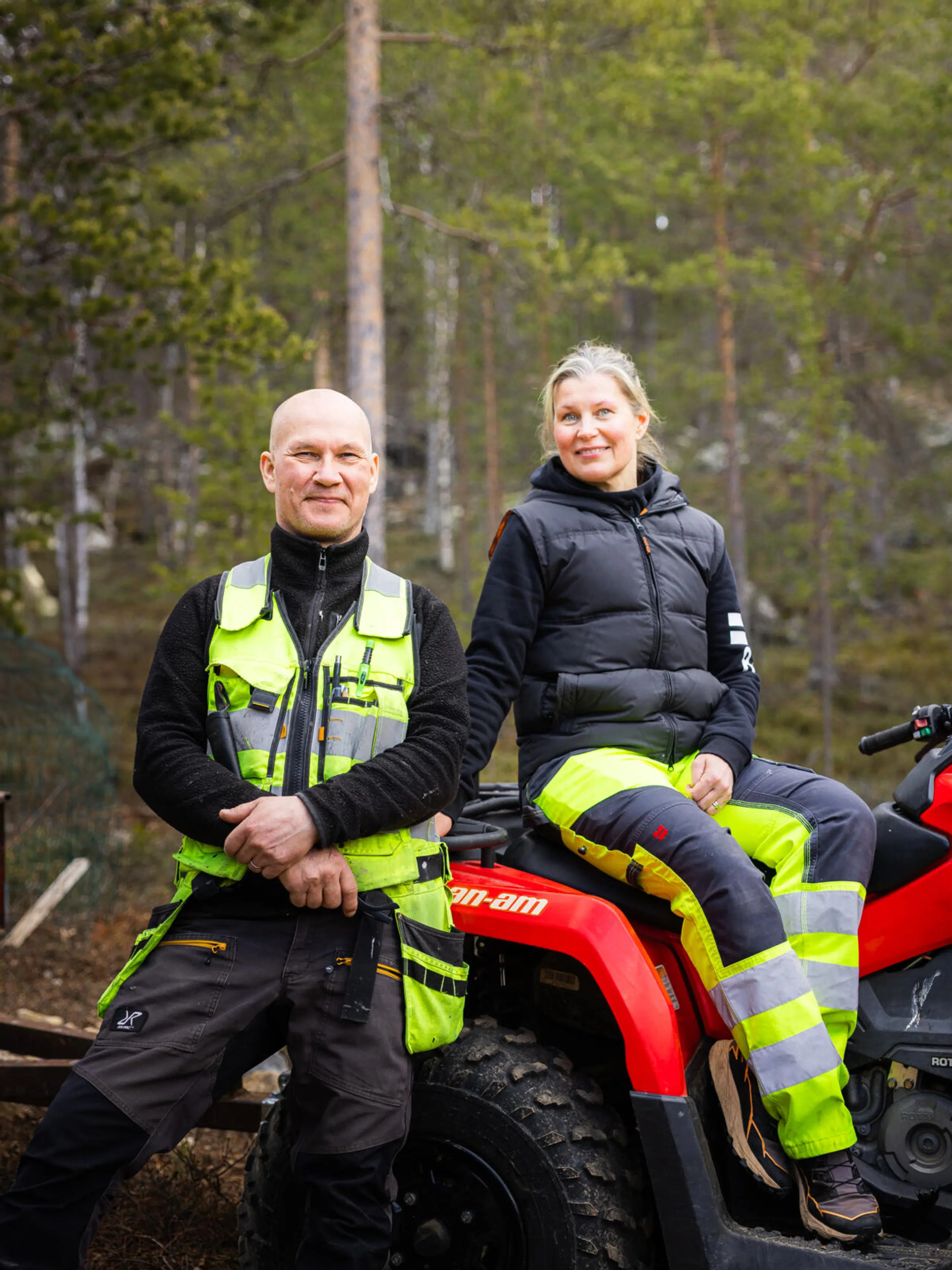 Kaksi henkilöä metsän edustalla, toinen seisoo ja toinen istuu punaisen mönkijän päällä.