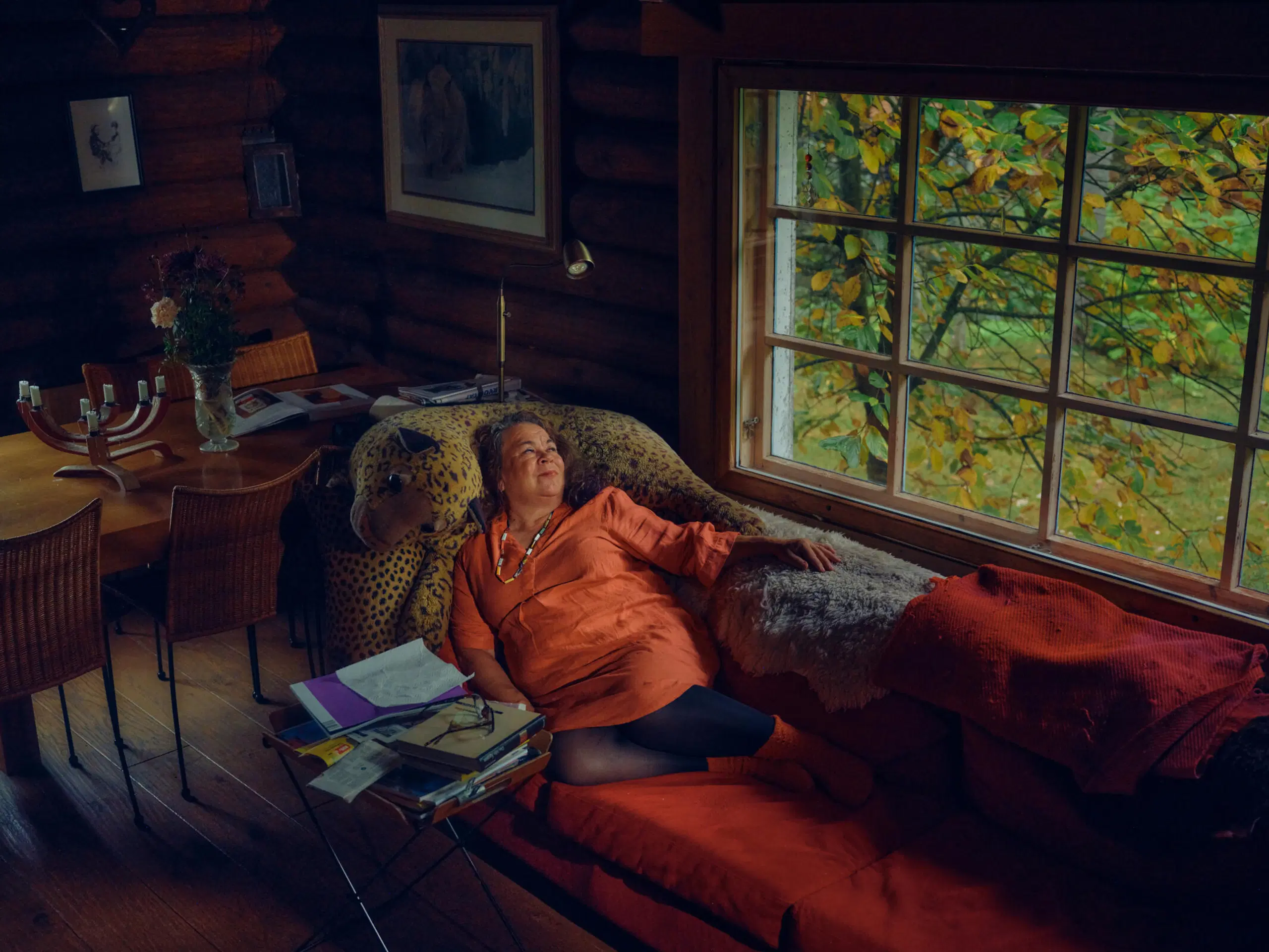 Henkilö lepää sohvalla hirsitalossa, syksyinen maisema ikkunasta, lämmin oranssi valaistus