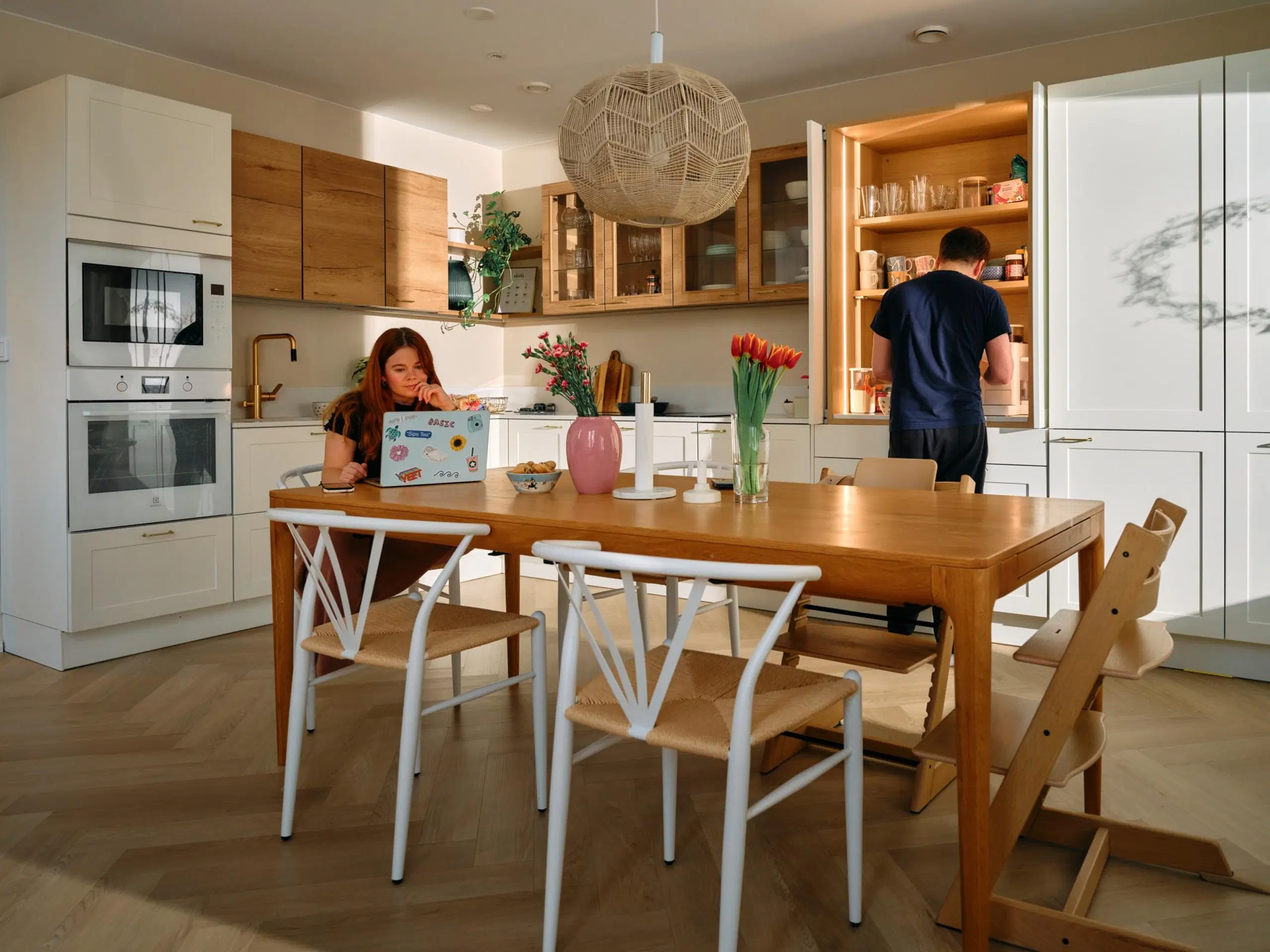 Salla Salmela istuu ruokapöydän ääressä kannettavan tietokoneen kanssa. Markus Salmela seisoo taustalla keittiössä.