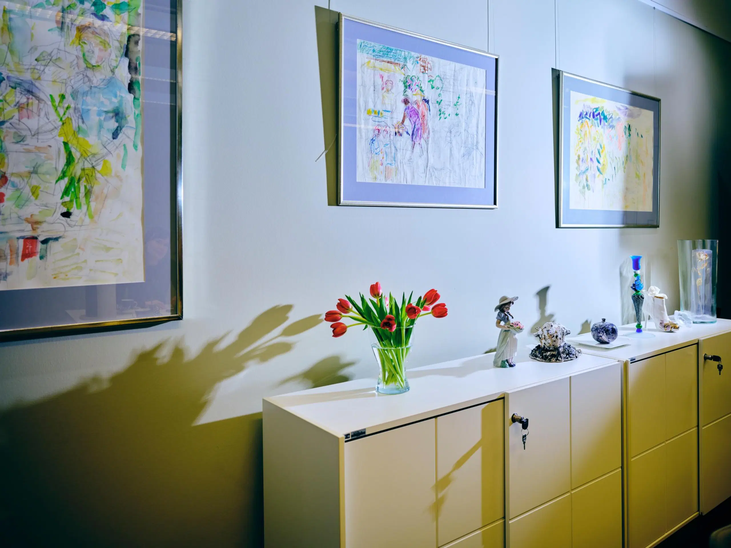 Valkoinen senkki, jonka päällä on tulppaanikimppu ja koriste-esineitä. Seinällä on kolme värikästä taulua.