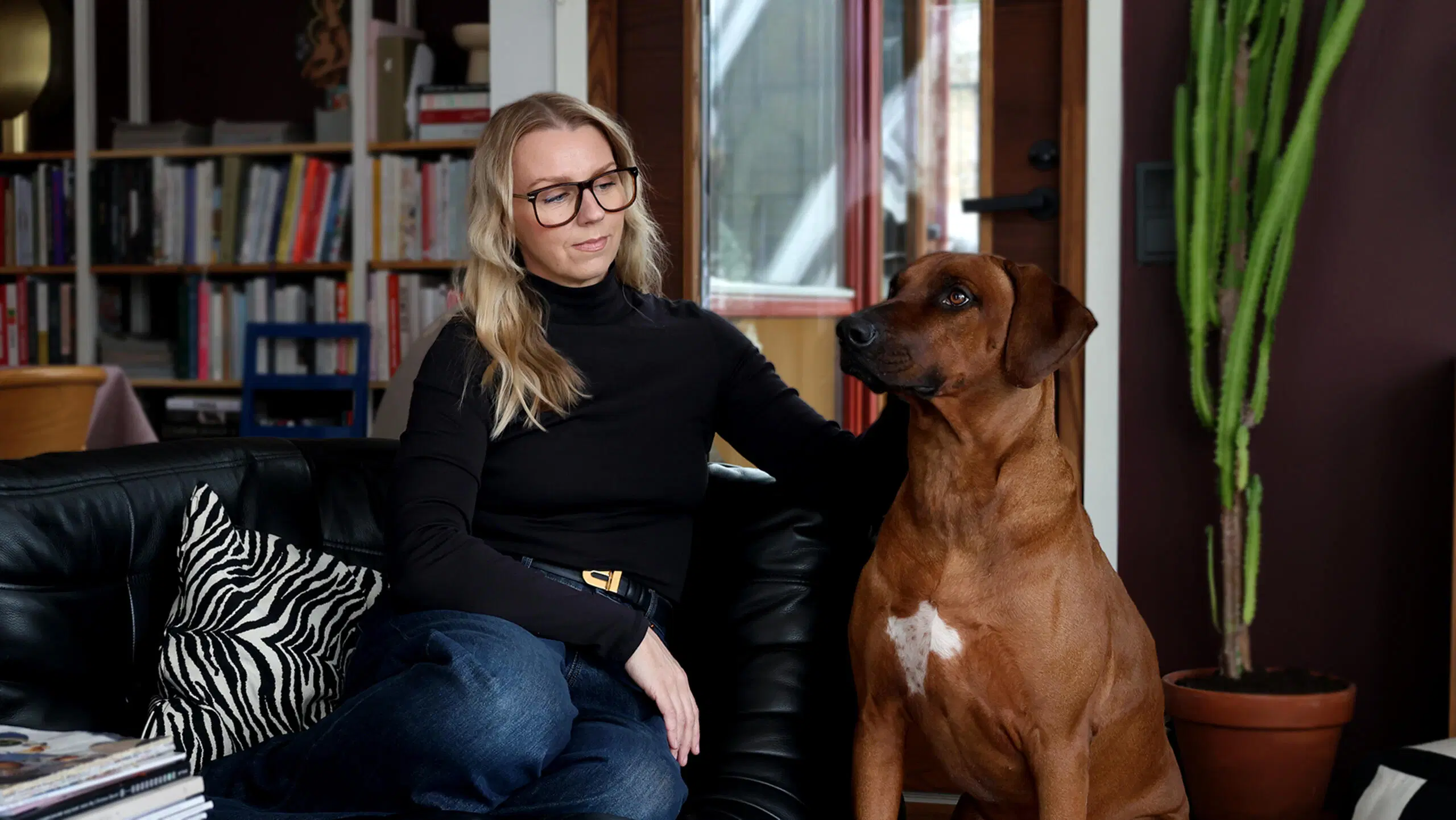 Hanna Himanka istuu mustalla sohvalla ja silittää isoa ruskeaa koiraa.