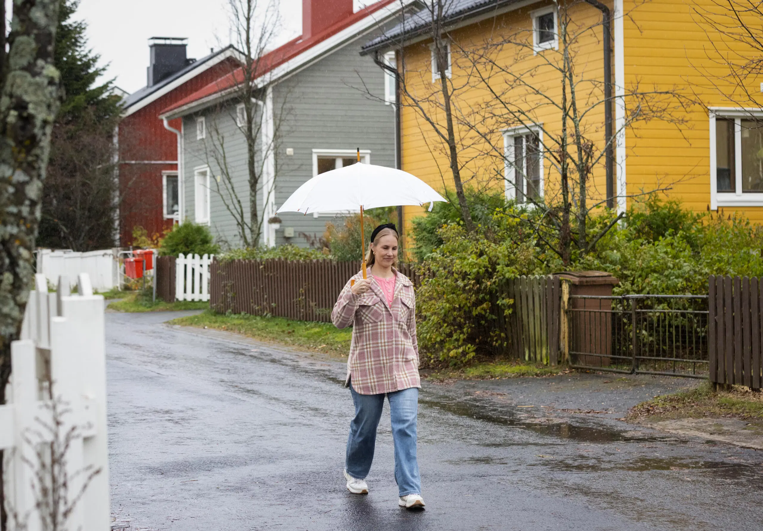 Nainen kävelee sateessa valkoinen sateenvarjo kädessä värikkäiden puutalojen kadulla.
