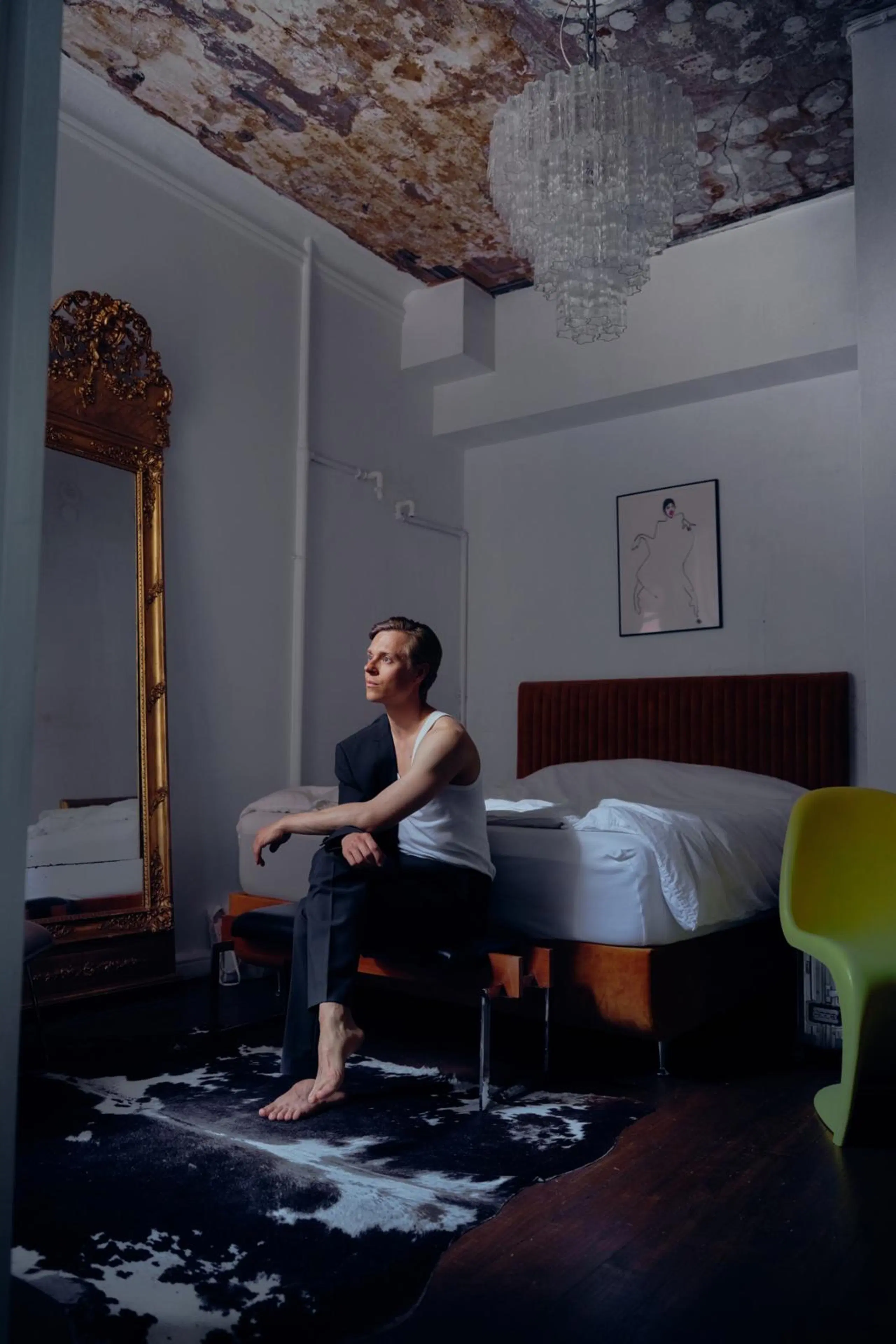 Atte Kilpinen istuu penkillä sängyn päädyssä. Huoneessa on myös kultakehyksinen kokovartalopeili, lasinen kattokruunu ja keltainen muovituoli.