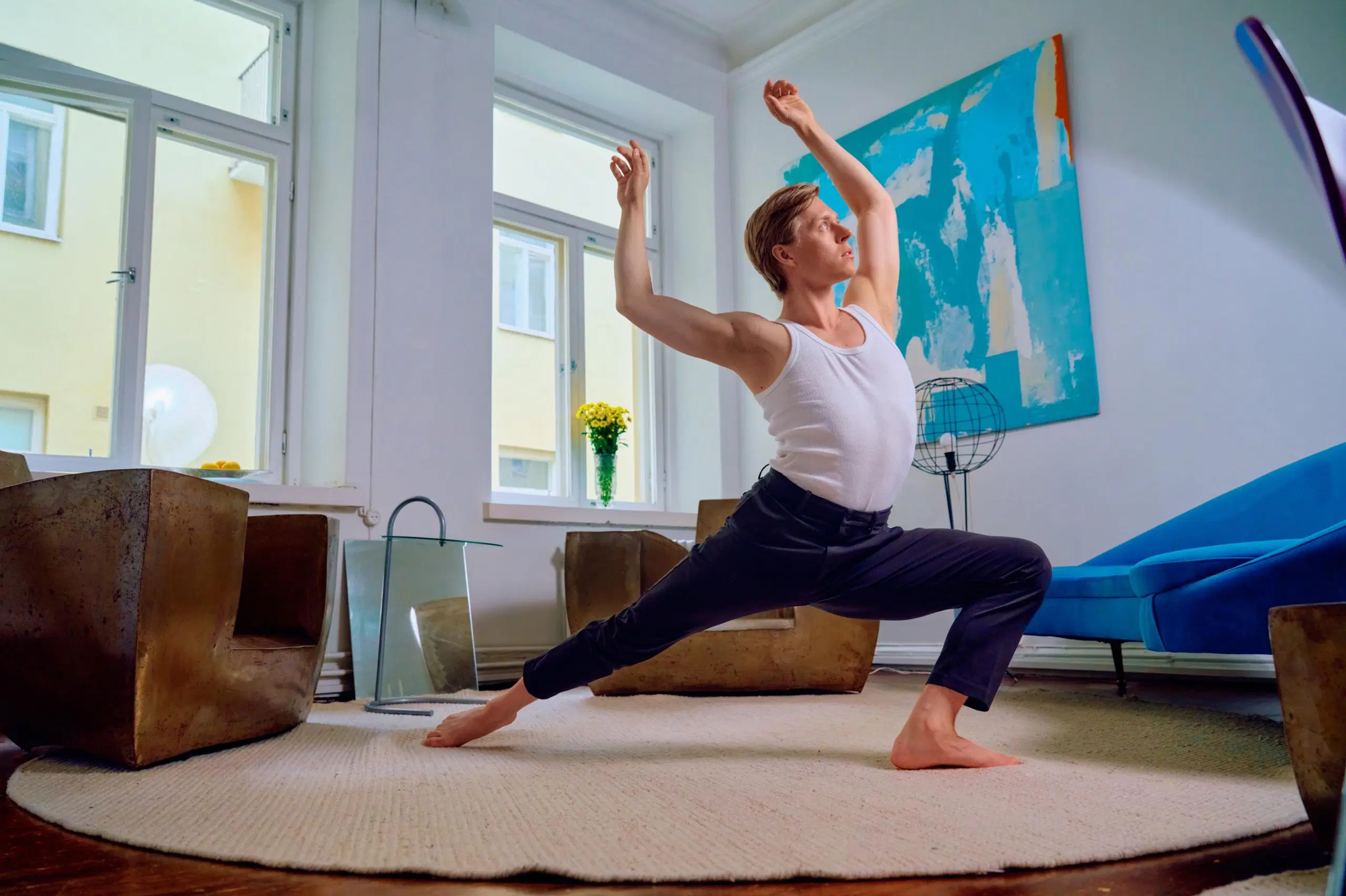 Atte Kilpinen venyttelee keskellä olohuonetta. Alla on iso pyöreä matto ja ympärillä kaksi rautanojatuolia sekä sininen sohva.