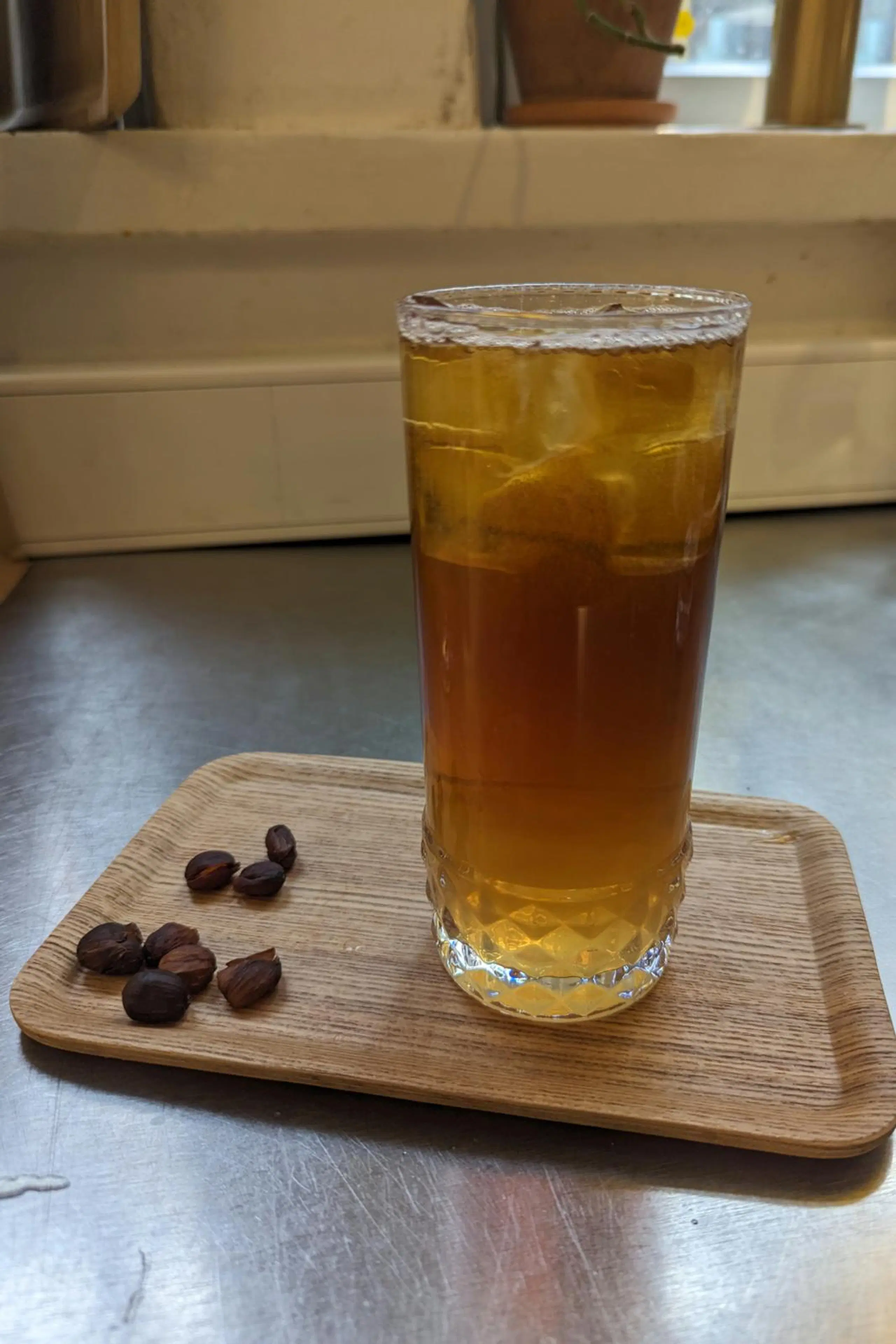 Vaaleanruskea juoma korkeassa lasissa. Lasi on puisella alustalla ja sen vieressä on muutamia kahvipapuja. 