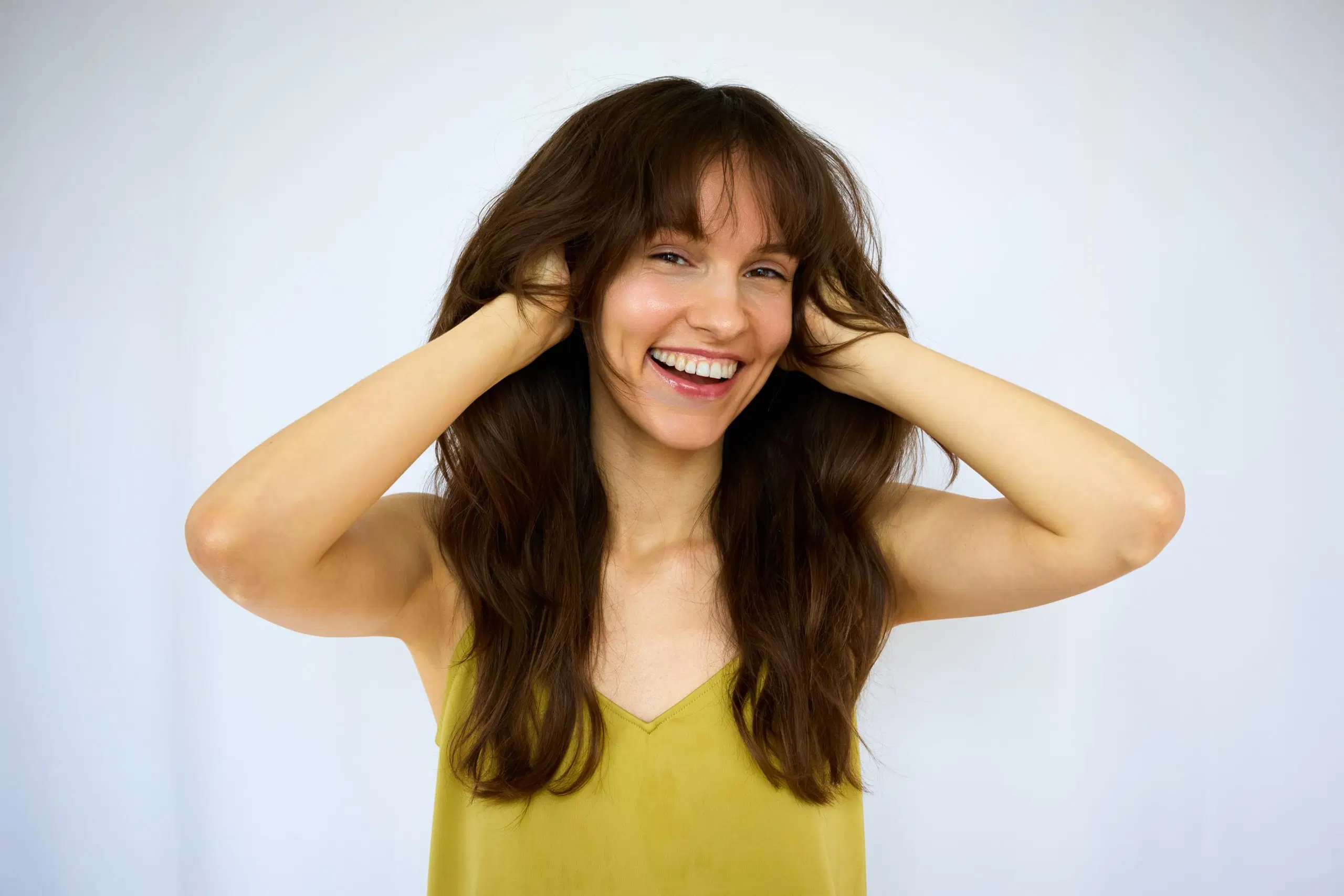 Iloisesti hymyilevä, tummahiuksinen nainen haroo hiuksiaan käsillään