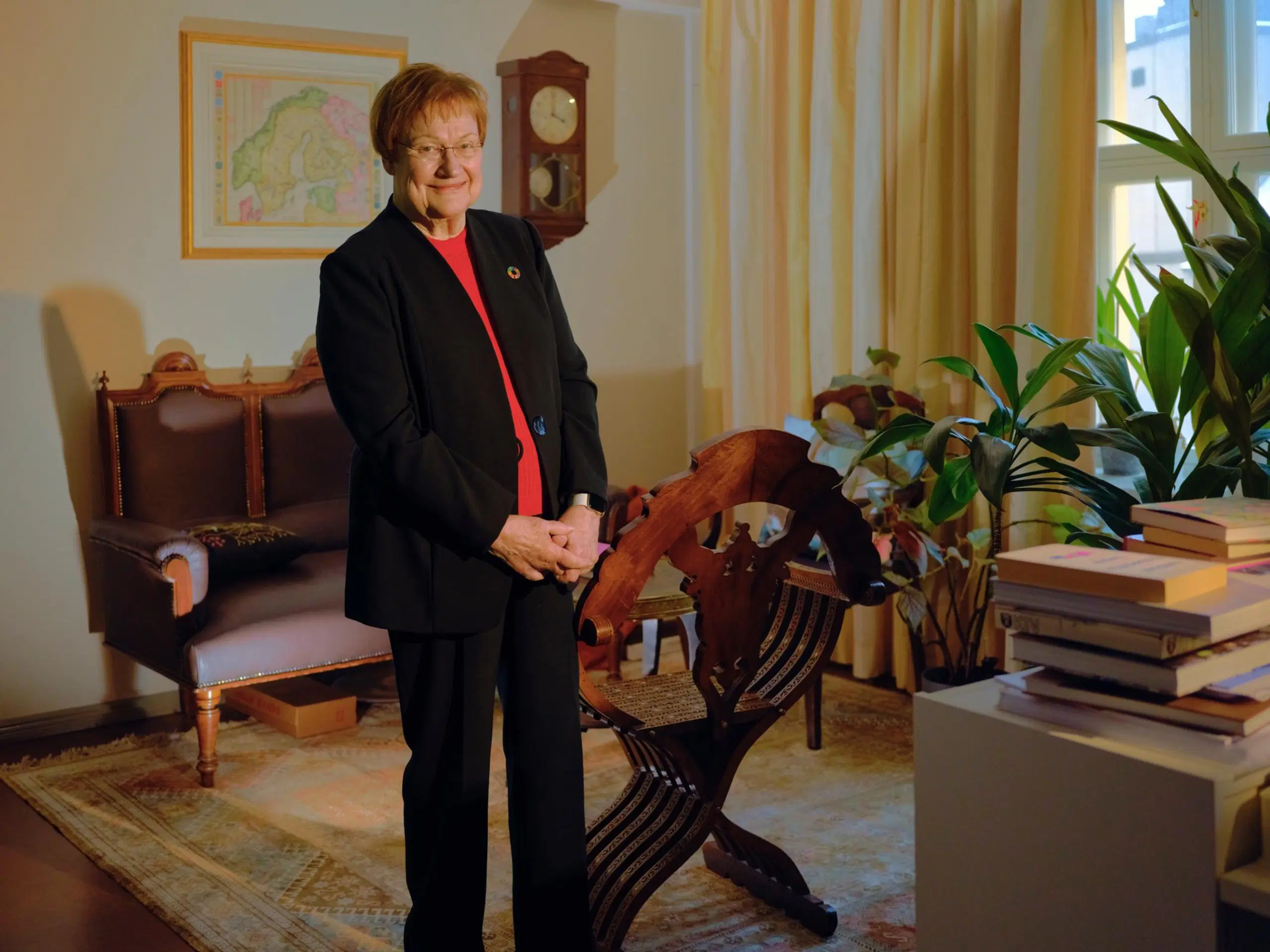 Tarja Halonen seisoo ja hymyilee. Taustalla antiikkikalusteita, vanha kartta ja seinäkello.