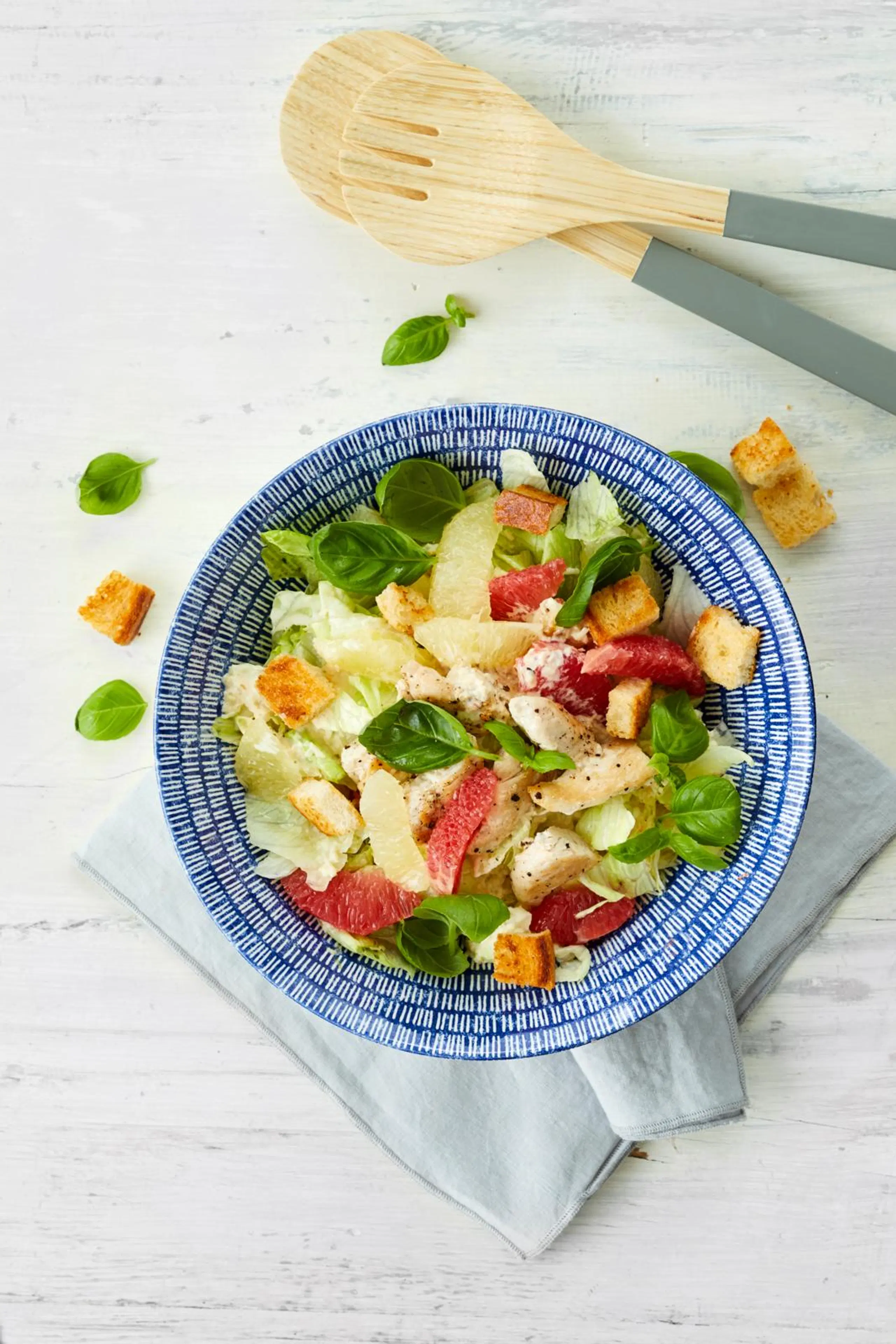 Greippi-kanacaesarsalaattia sinisellä lautasella, vieressä puiset ottimet