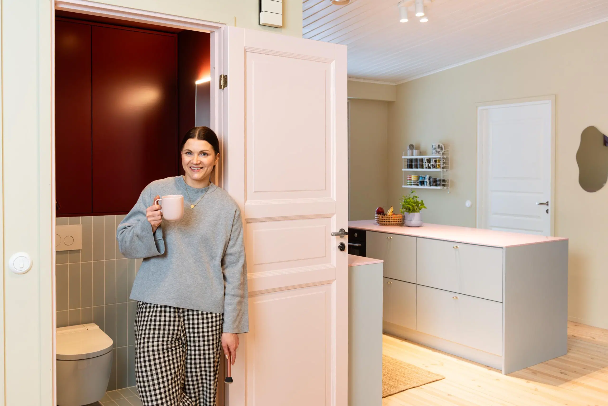 Maru Hautala nojailee vessan ovenpieleen käsissään kahvikuppi ja meikkisuti. Taustalla näkyy myös keittiö.