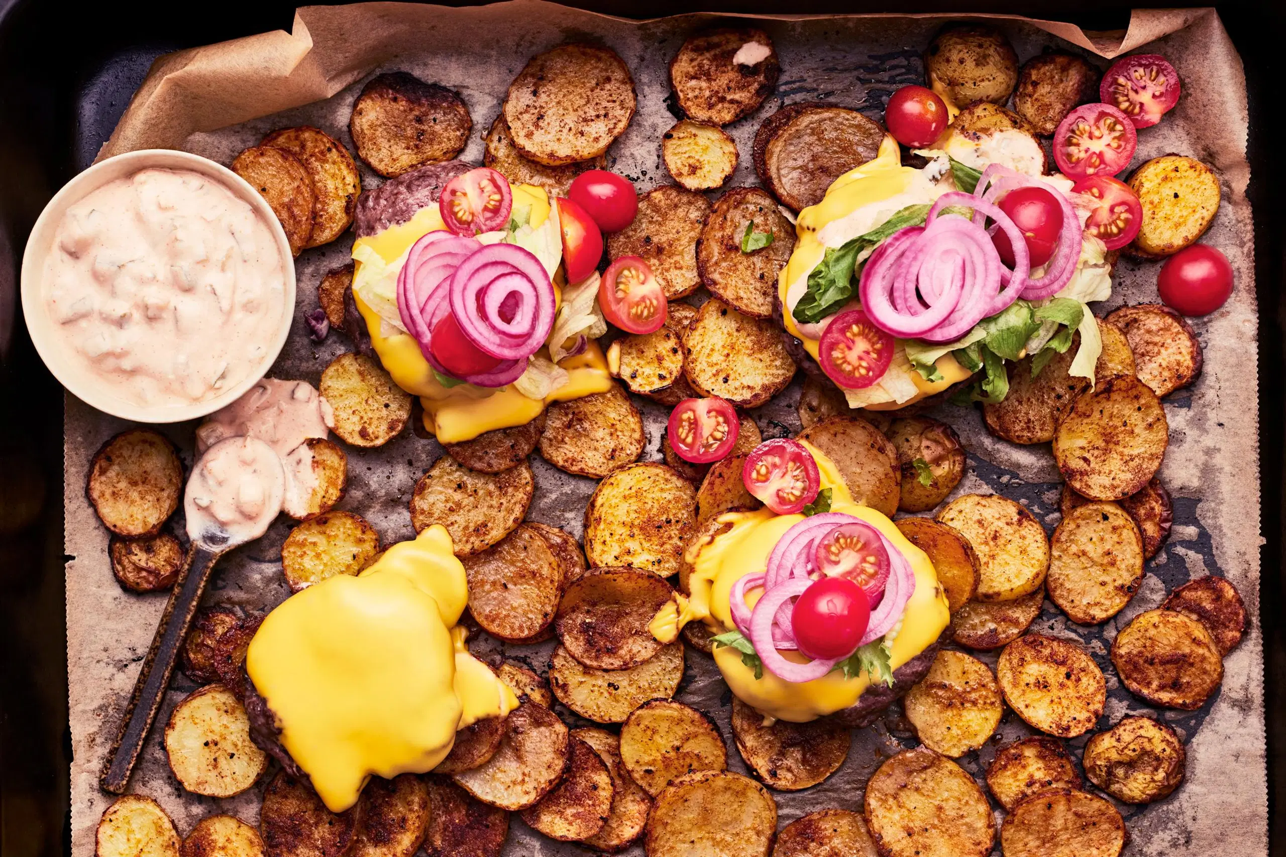 Hampurilaispihvejä, perunaviipaleita ja hampurilaiskastiketta pellillä.