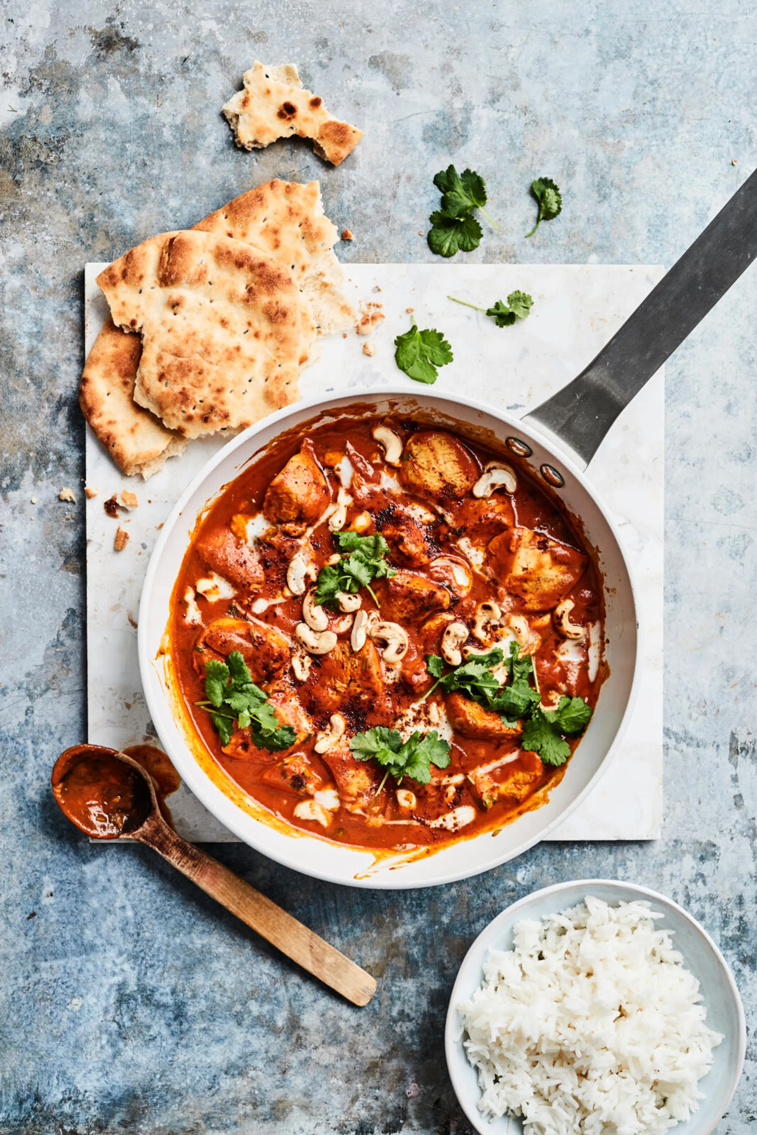 Kanaa punaisessa kastikkeessa pannulla. Päällä korianteria ja cashew-pähkinöitä, vieressä naan-leipää ja riisiä.