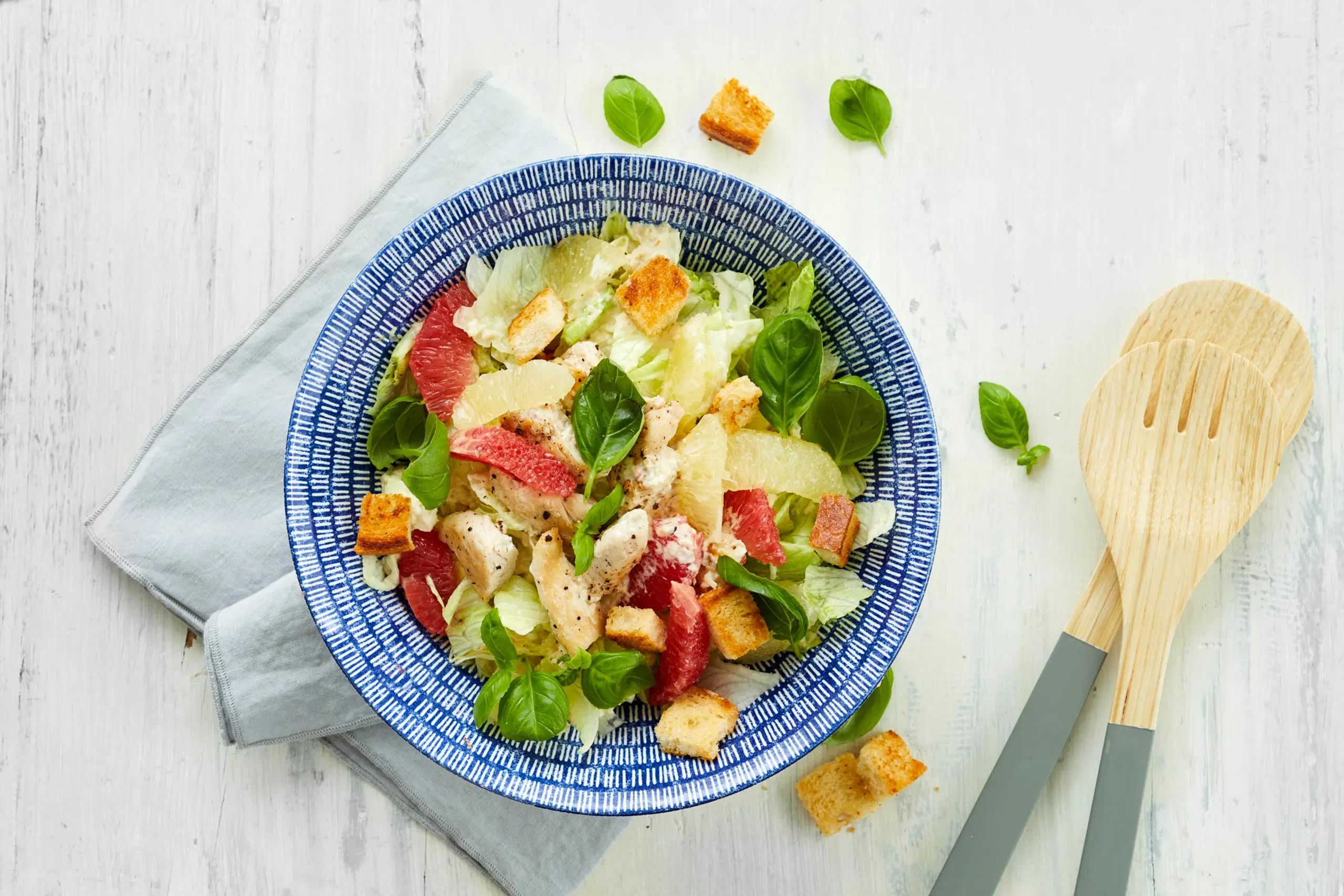 Greippi-kanacaesarsalaattia sinisellä lautasella, vieressä puiset ottimet