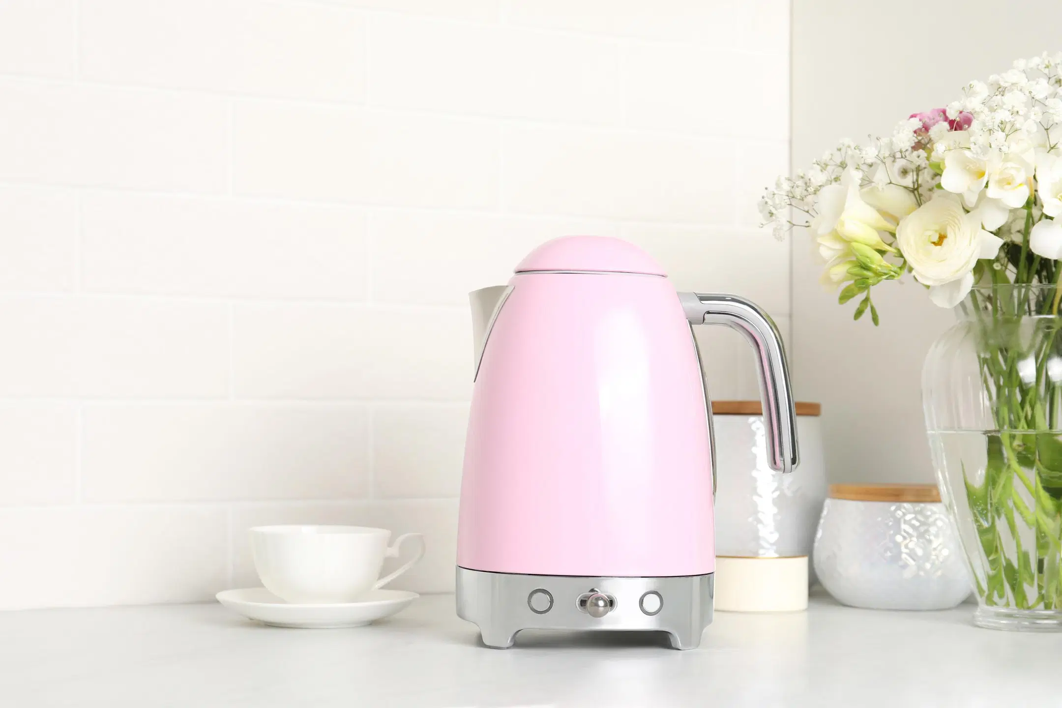 Vaaleanpunainen vedenkeitin, valkoinen kahvikuppi, kaksi purkkia ja kukkia maljakossa keittiötasolla.