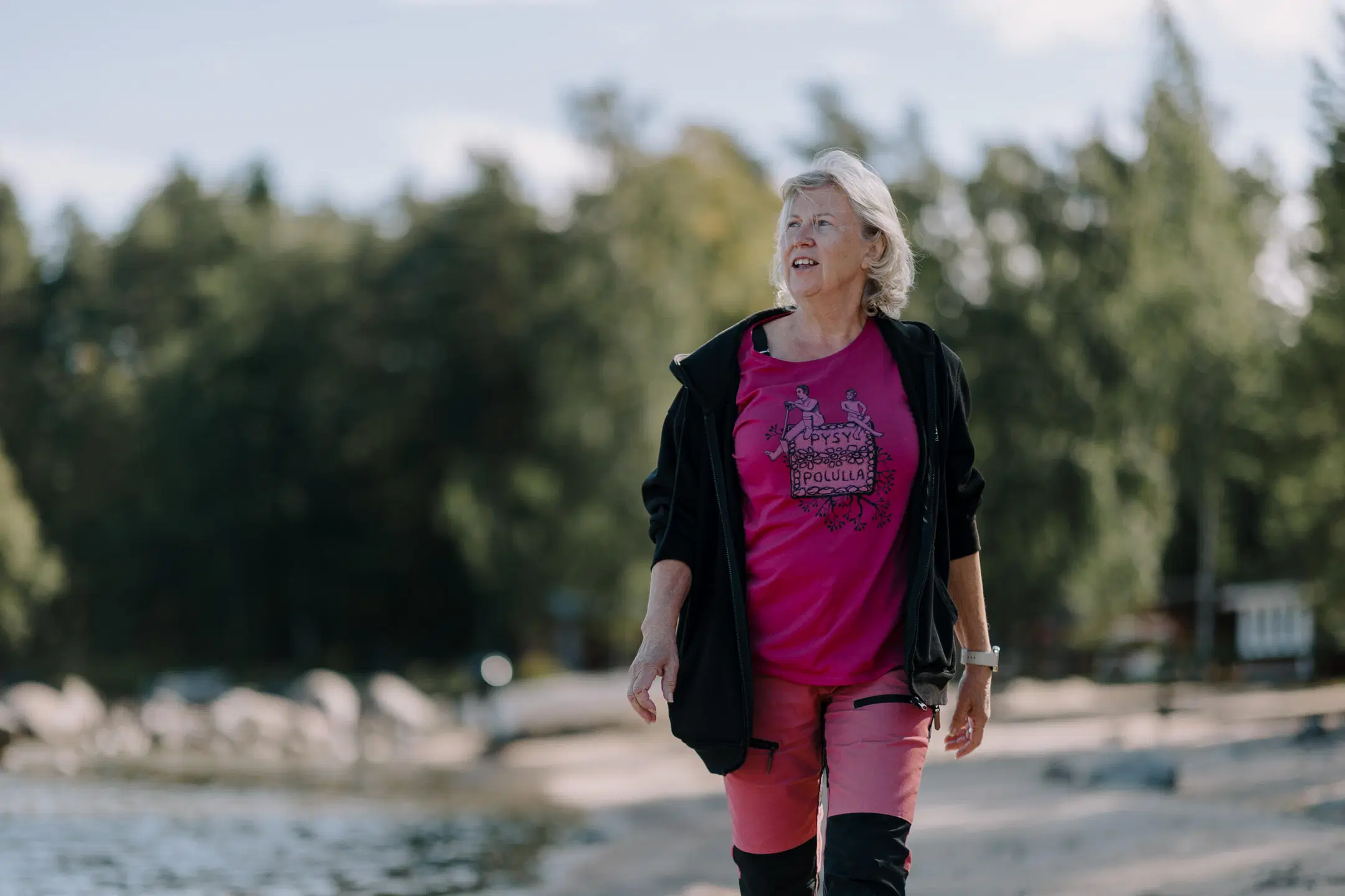 Nainen kävelee rannalla metsän edessä, yllään pinkki paita ja musta takki, aurinkoinen päivä.