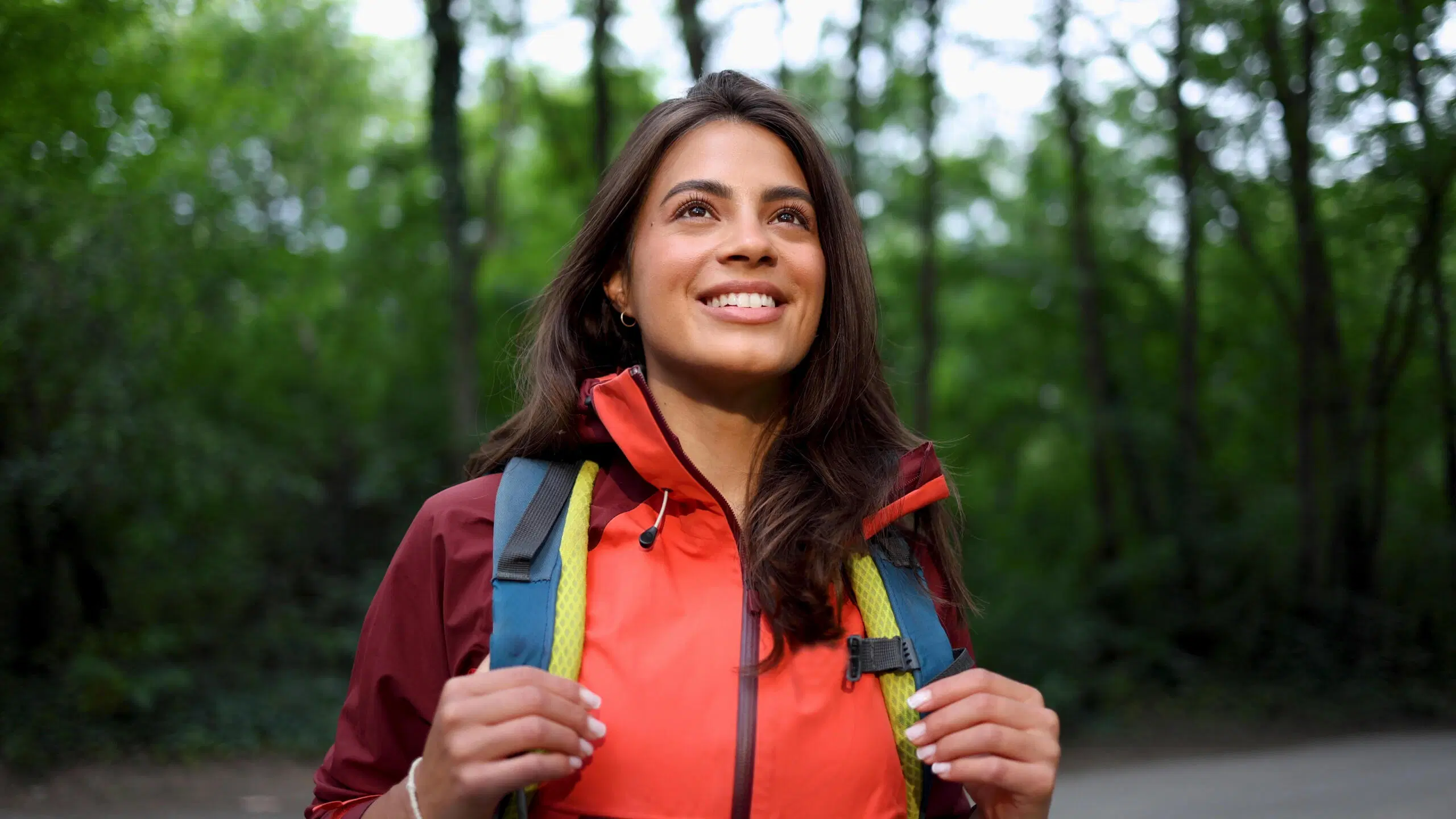 Hymyilevä nainen punaisessa ulkoilutakissa ja reppu selässä