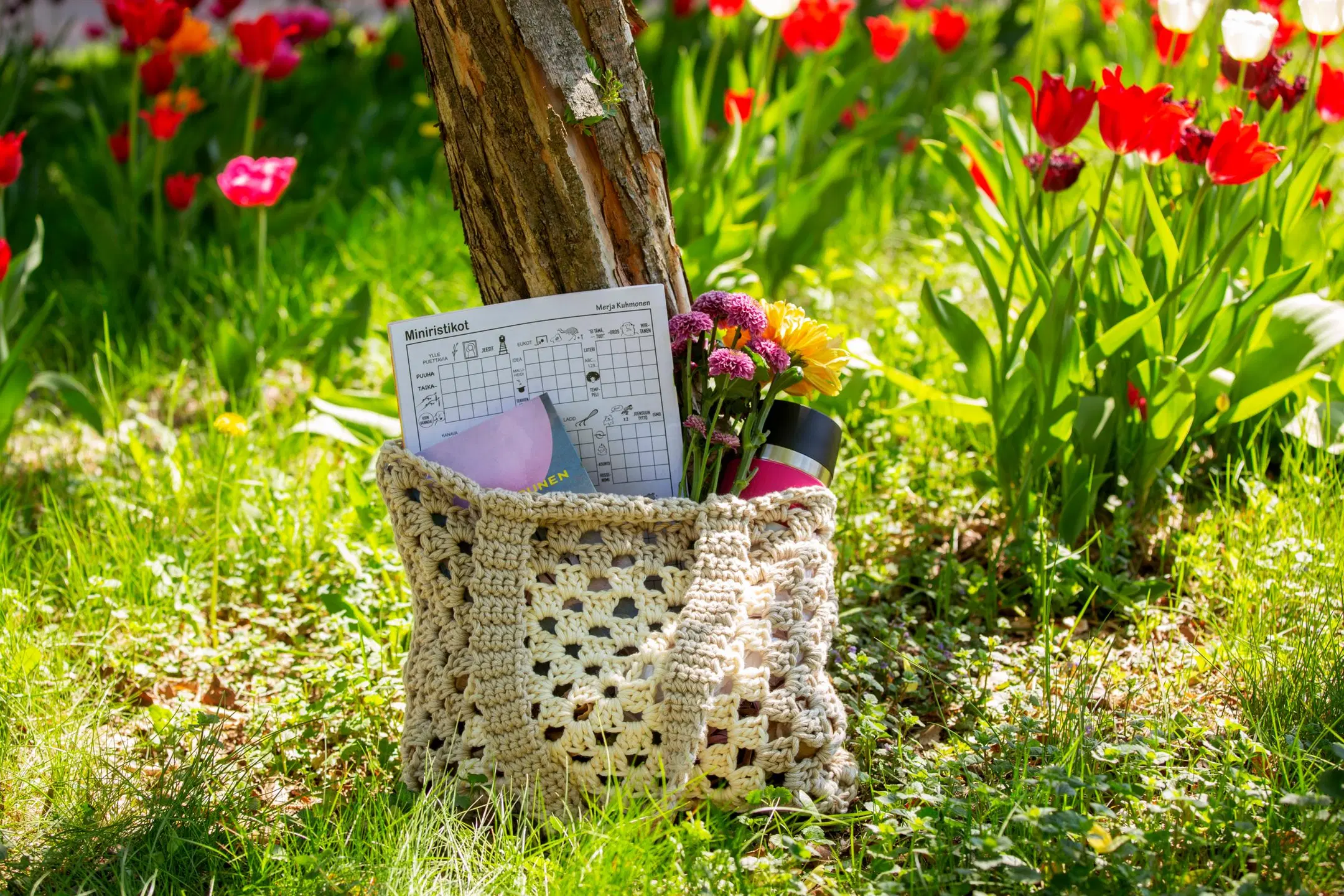 Isoäidinneliöistä tehty kassi, jonka sisällä ristikkolehti, kirja, kukkia ja juomapullo