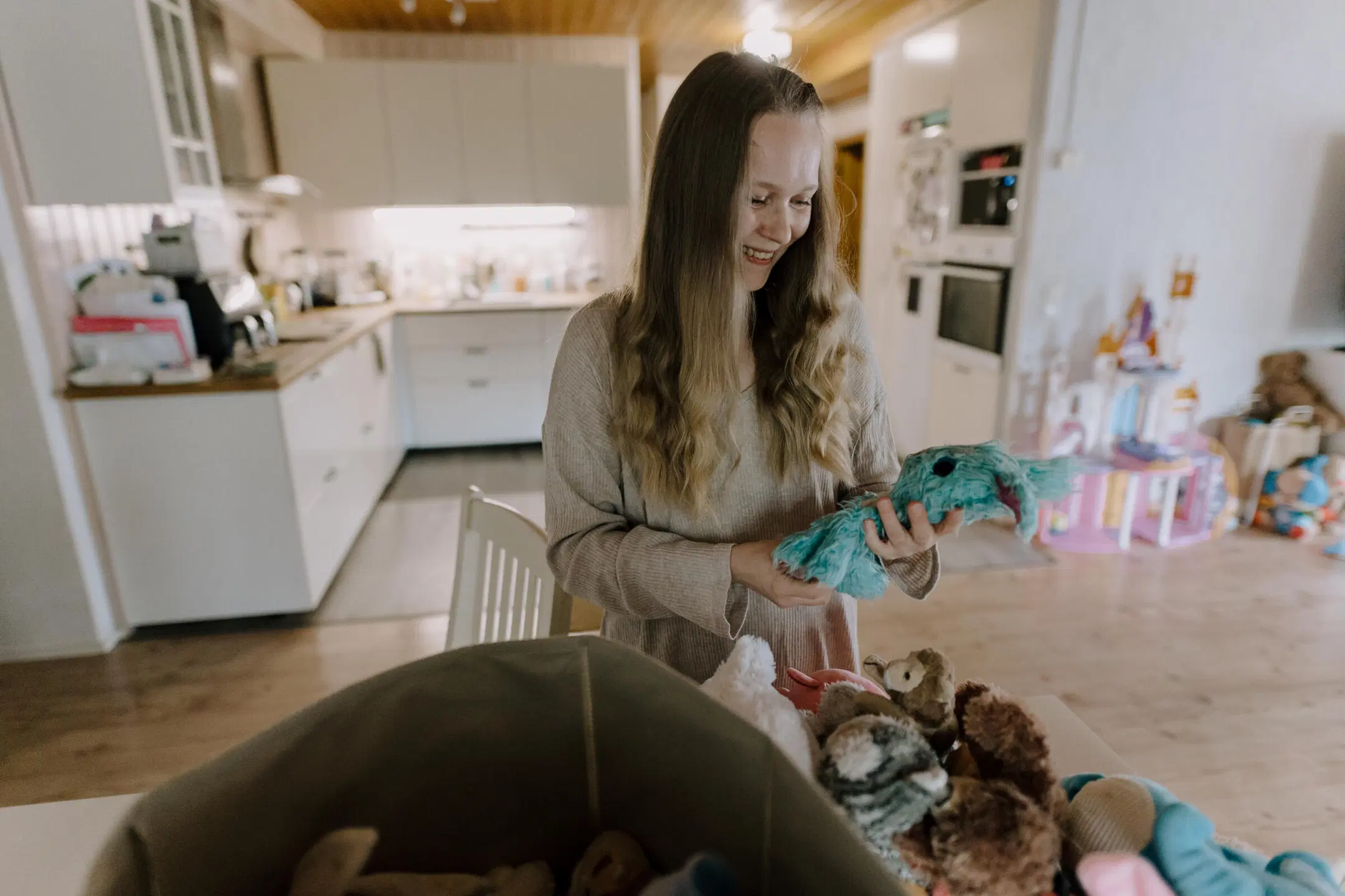 Hymyilevä nainen olohuoneessa pitelee siniturkoosia pehmolelua, leluja edessä korissa.